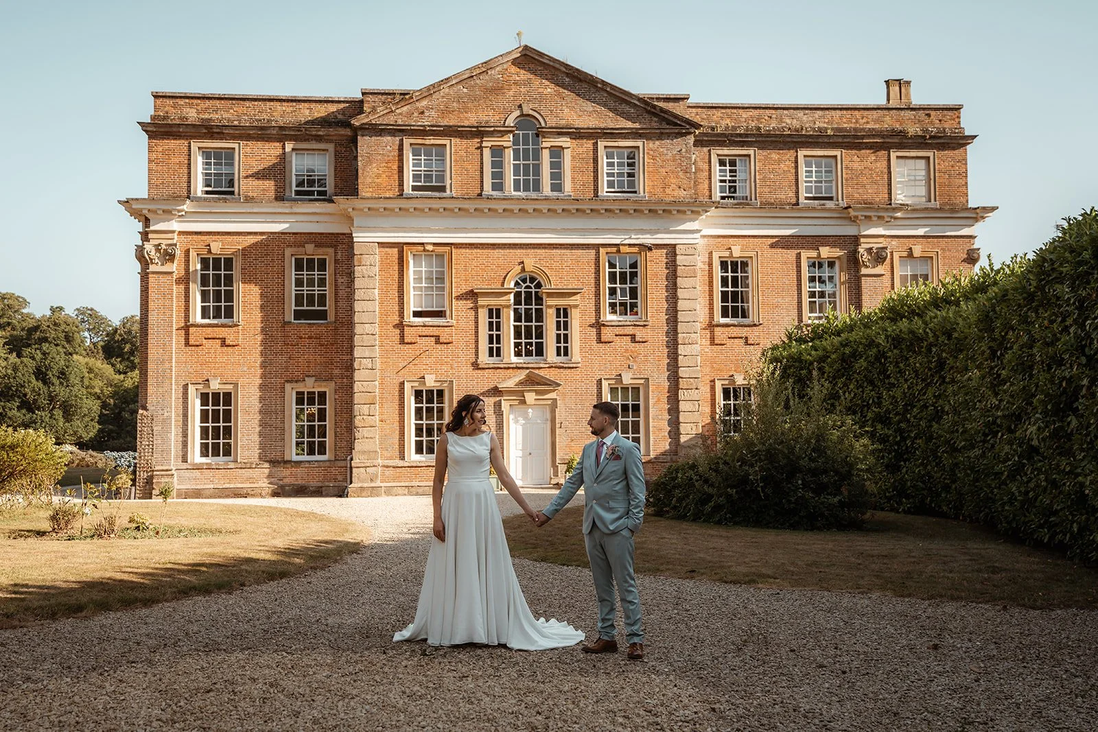 Michelle & Andy hold hands during relaxed wedding portraits at Crowcombe Court in Somerset, UK, standing in front of the historic brick manor as soft evening light highlights the bride’s flowing dress and the groom’s tailored suit.