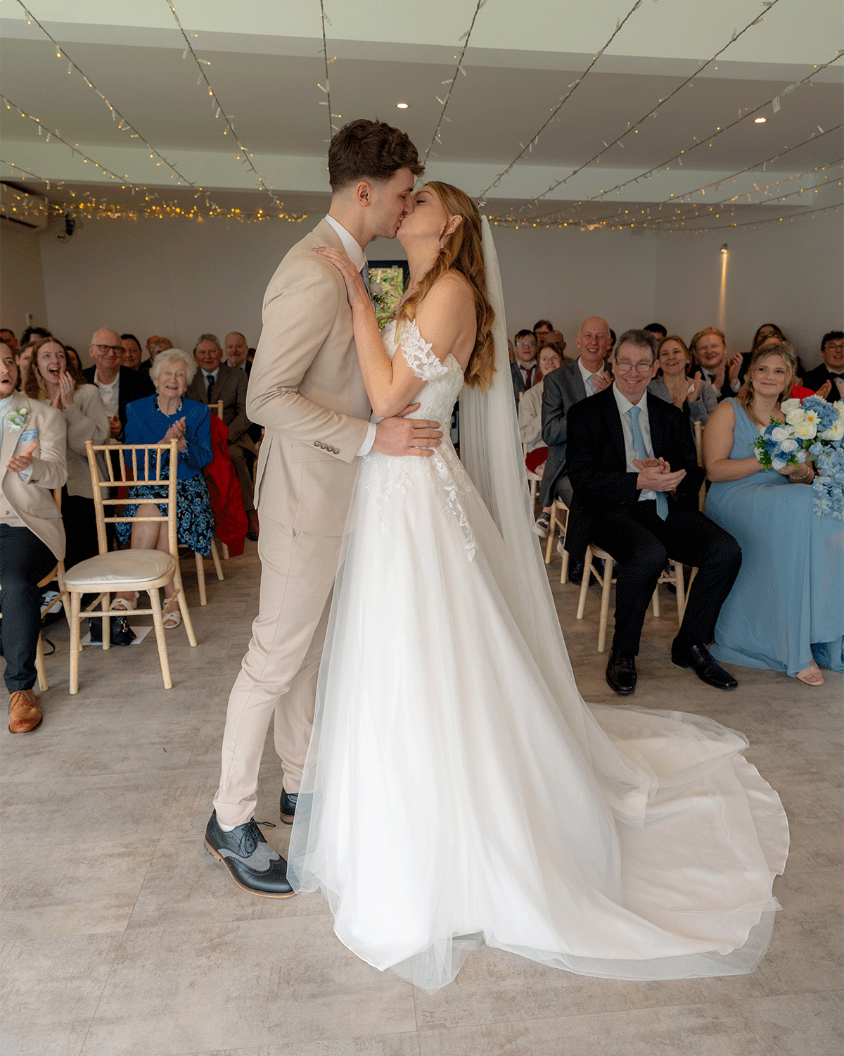 A bride and groom sharing their first kiss during a wedding ceremony, with friends and family seated and clapping in the background.