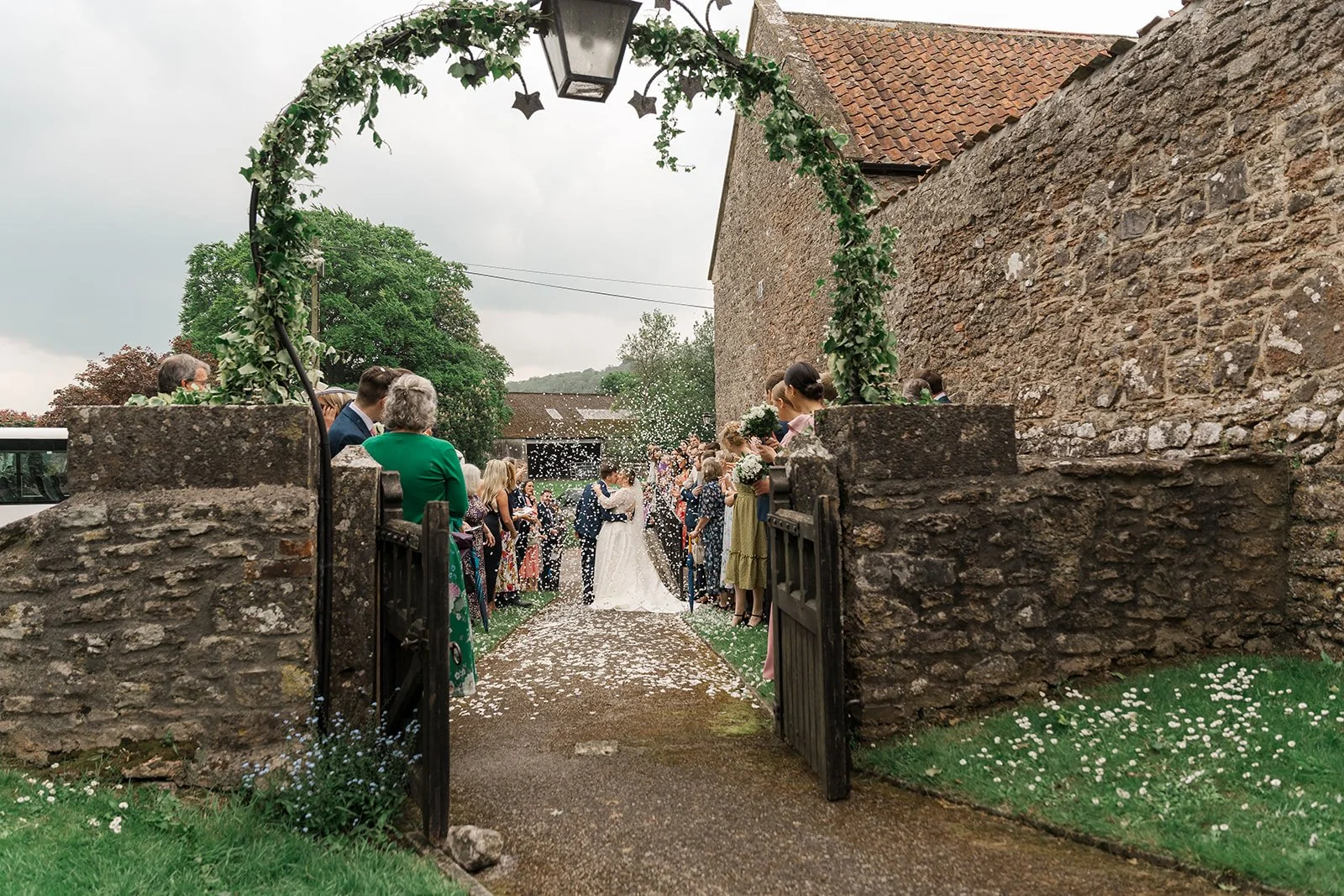 Beth & Tom share a confetti-filled wedding exit at Bishop’s Palace in Somerset, UK, framed by a stone gate and leafy archway as guests line the path and celebrate around them, creating a romantic outdoor wedding moment.