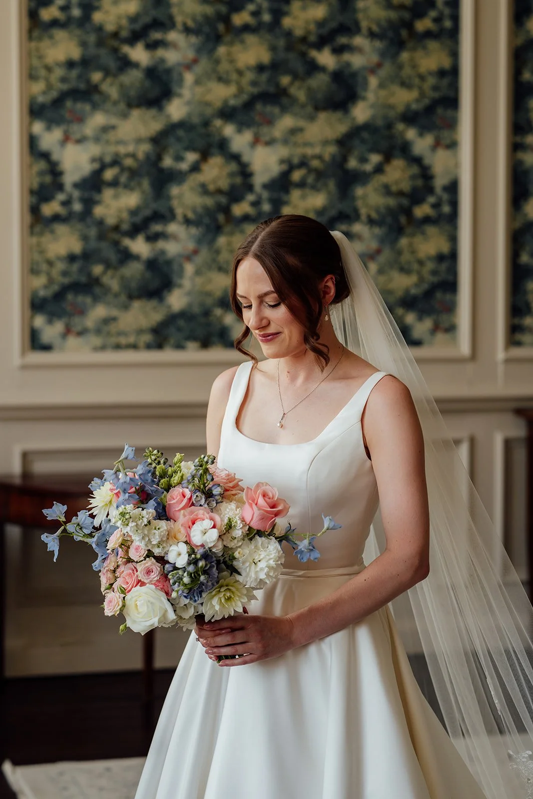 Rachel stands indoors at Brympton House in Somerset holding her pastel pink, blue and white wedding bouquet while wearing her bridal gown and veil, photographed by Just James Weddings.