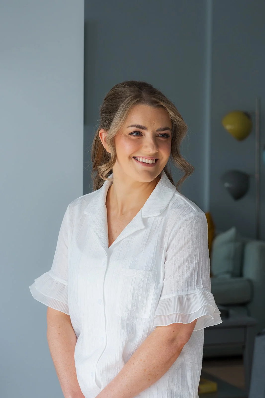 Anna smiles in a white bridal getting-ready blouse by a window before Anna and Tom’s Winkworth Farm wedding in the Cotswolds, UK. This natural bridal portrait captures soft light, elegant hair and makeup, and relaxed wedding morning photography.