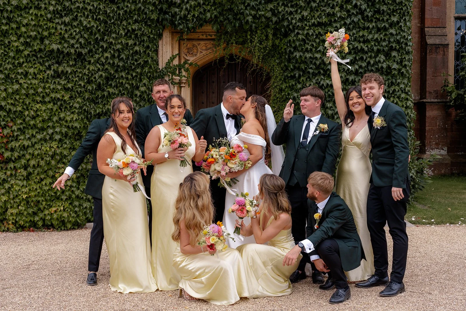 Leacarla &amp; Carl at St. Audries Park surrounded by their wedding party, captured in front of an ivy-covered wall as everyone smiles, poses and celebrates. The group holds bright floral bouquets while the couple share a kiss at the centre.