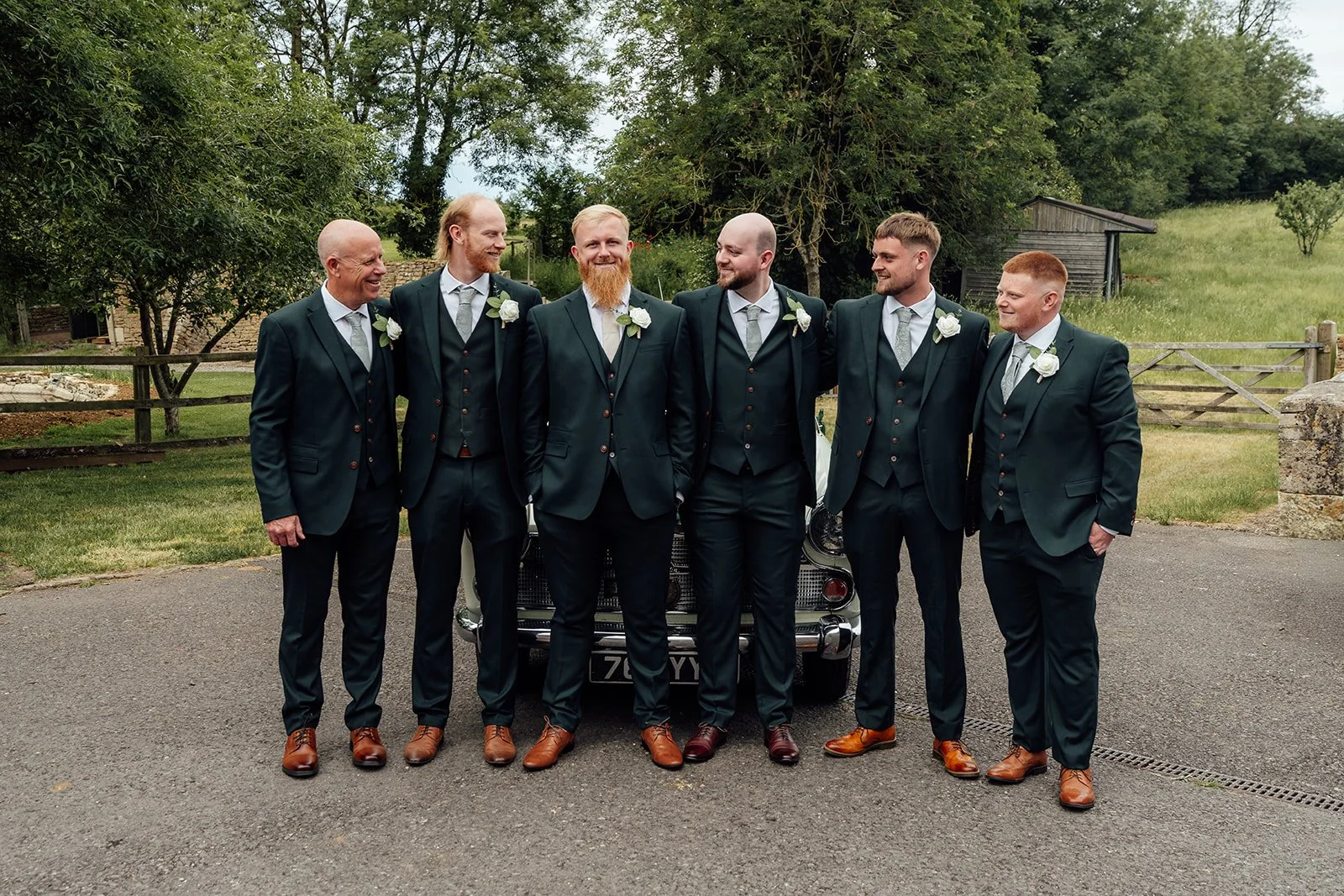 Relaxed groomsmen portrait for Louise & Jon’s wedding at Wick Farm Bath in Somerset, UK, with dark green suits, white buttonholes, brown shoes, a vintage car, countryside gates, and natural laughter before the ceremony.