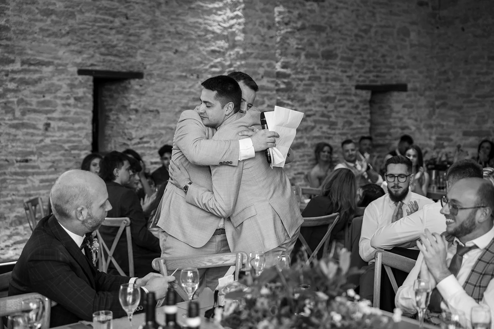 Black and white photo of Sam & Gavin hugging during speeches at The Great Barn at Aynho in the Cotswolds, photographed by Just James Weddings for emotional reception moments.