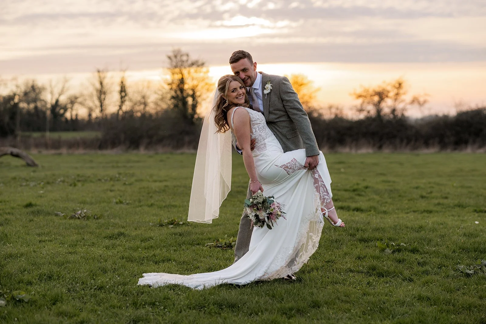 Anna and Tom share a joyful and playful moment on the open green fields of Winkworth Farm in Cotswolds at golden hour, with a warm winter sunset glowing behind them as Tom dips Anna, her lace train and veil sweeping across the grass.
