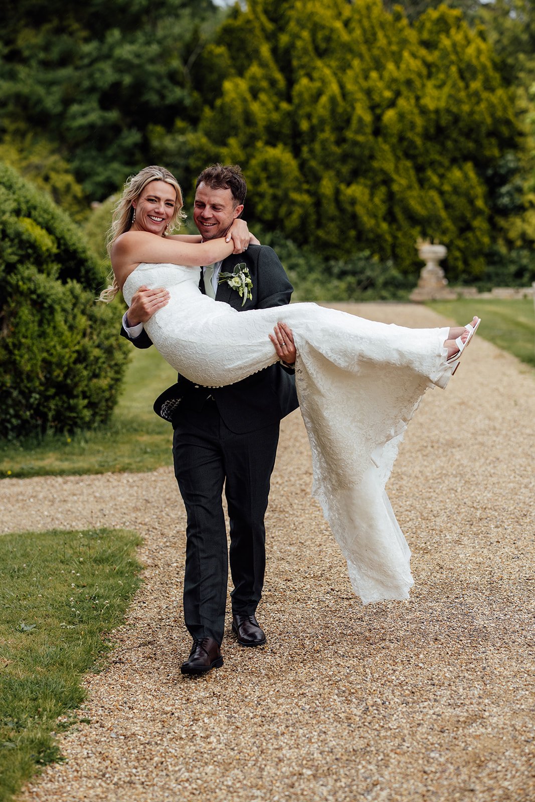 Amy & Matt laugh as the groom carries the bride along a gravel path at Orchardleigh Estate in Somerset, with green gardens around them and her lace dress flowing, captured by Just James Weddings.