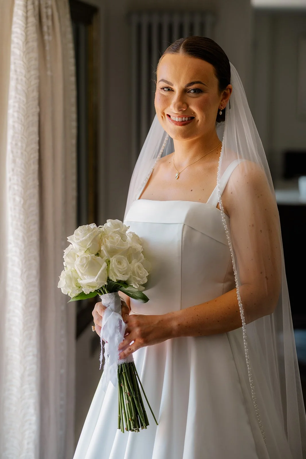 Bridal portrait of Lydia holding a white rose bouquet in her wedding dress and veil beside soft window light at Hencote Estate in Shropshire, UK, during Lydia & Kieran's elegant countryside wedding day, with classic details and a calm smile.