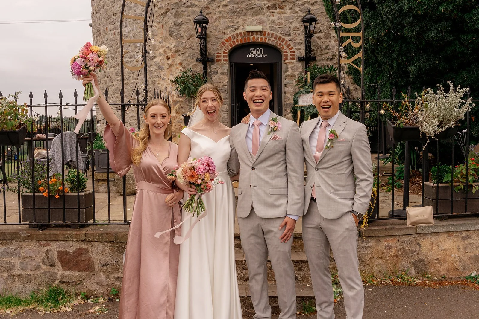 Brian & Josie celebrating with their wedding party outside Clifton Observatory in Somerset, holding bouquets and smiling together, photographed by Just James Weddings.