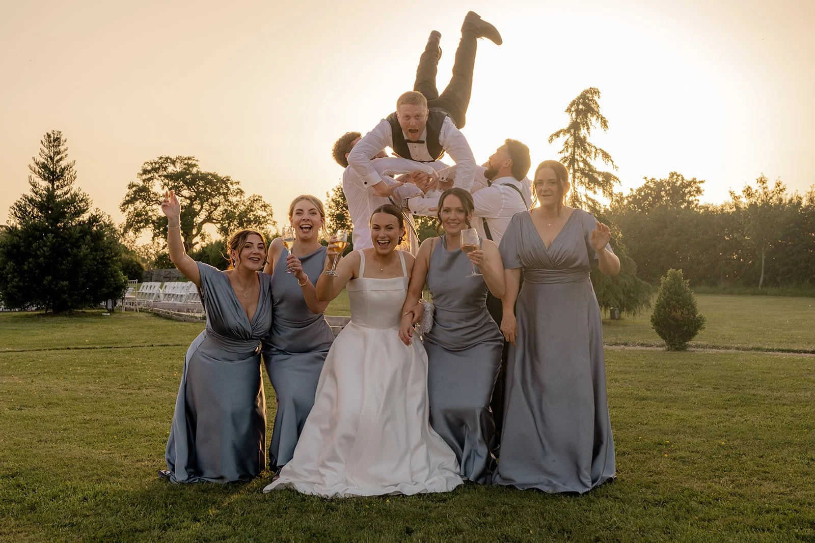Wedding party photo of Lydia & Kieran with bridesmaids and groomsmen on the lawn at Hencote Estate in Shropshire, UK, with the groom playfully lifted behind them during golden hour and everyone laughing in warm summer evening light together.
