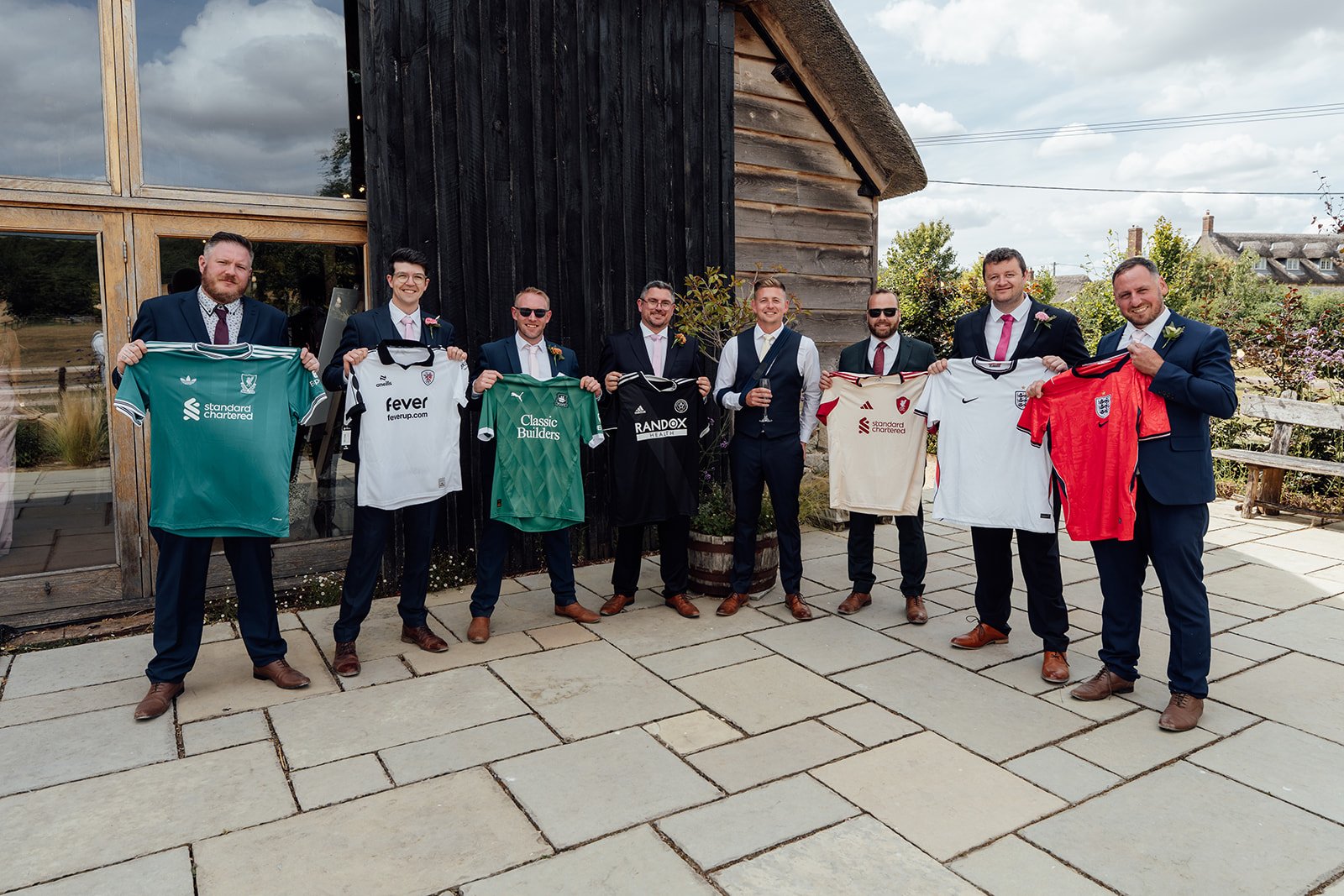 Tom and his groomsmen holding cricket shirts during a fun wedding party photo outside Bowerchalke Barn in Wiltshire, photographed by Just James Weddings.