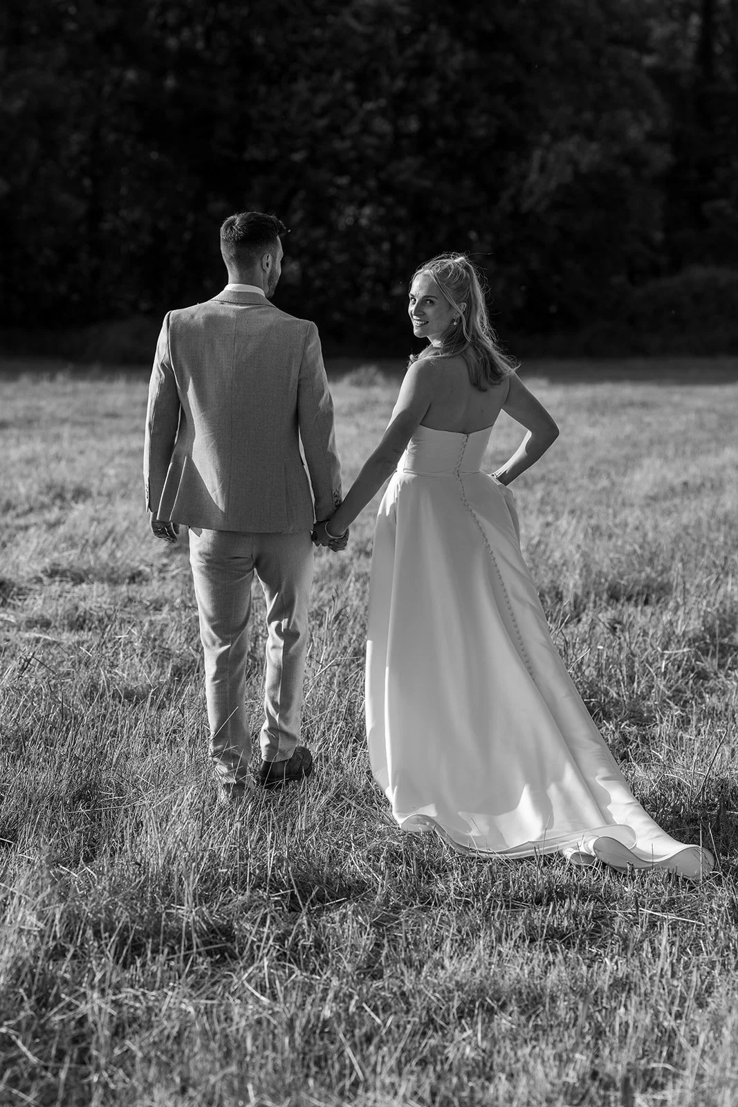 Black and white portrait of Louise & Christian walking hand in hand through a field at The Cider Barn in Somerset, with the bride looking back towards the camera, photographed by Just James Weddings.