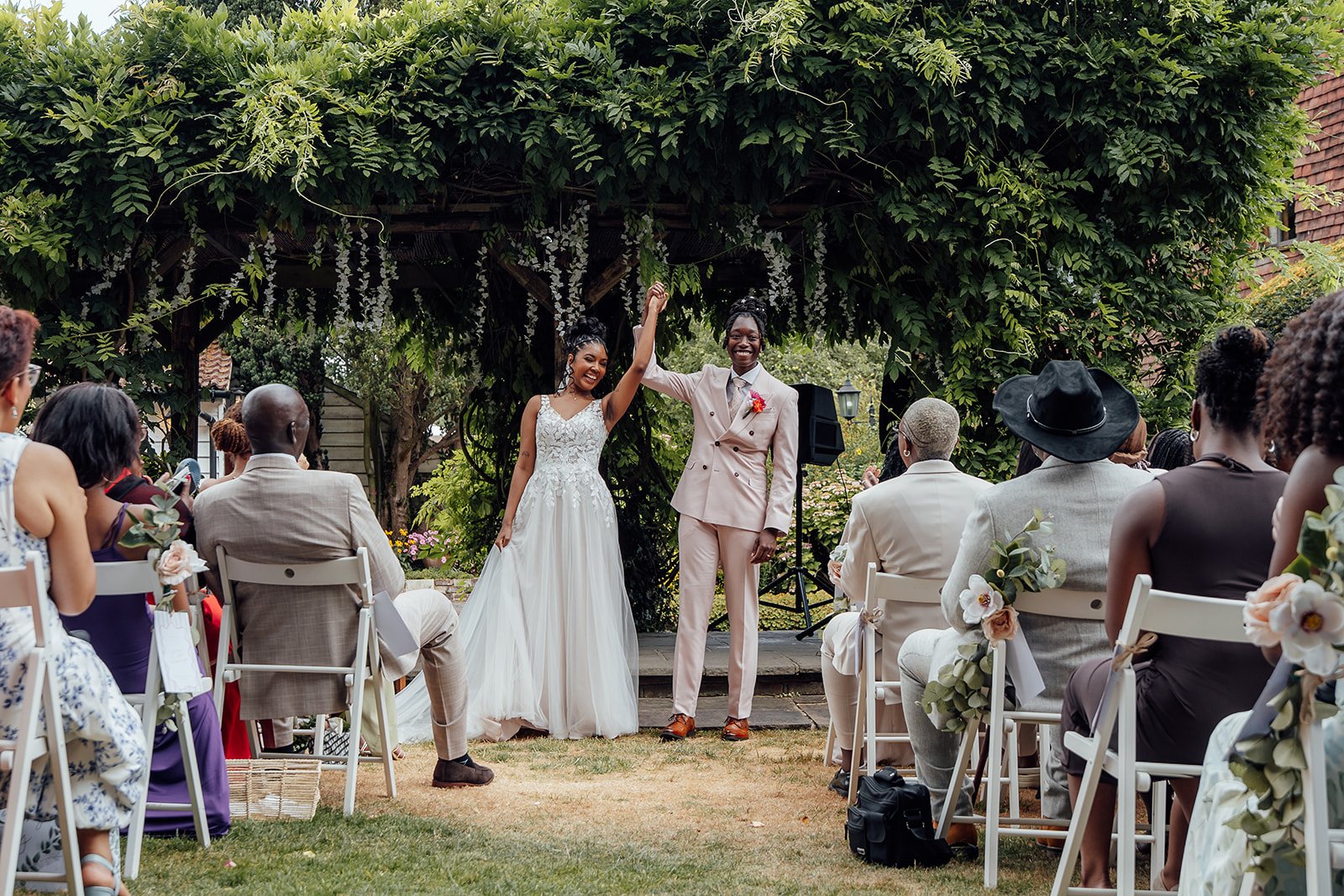 Kasey and Larisha smiling and raising their joined hands after their outdoor wedding ceremony at Oaks Farm Weddings in Surrey, surrounded by cheering guests.
