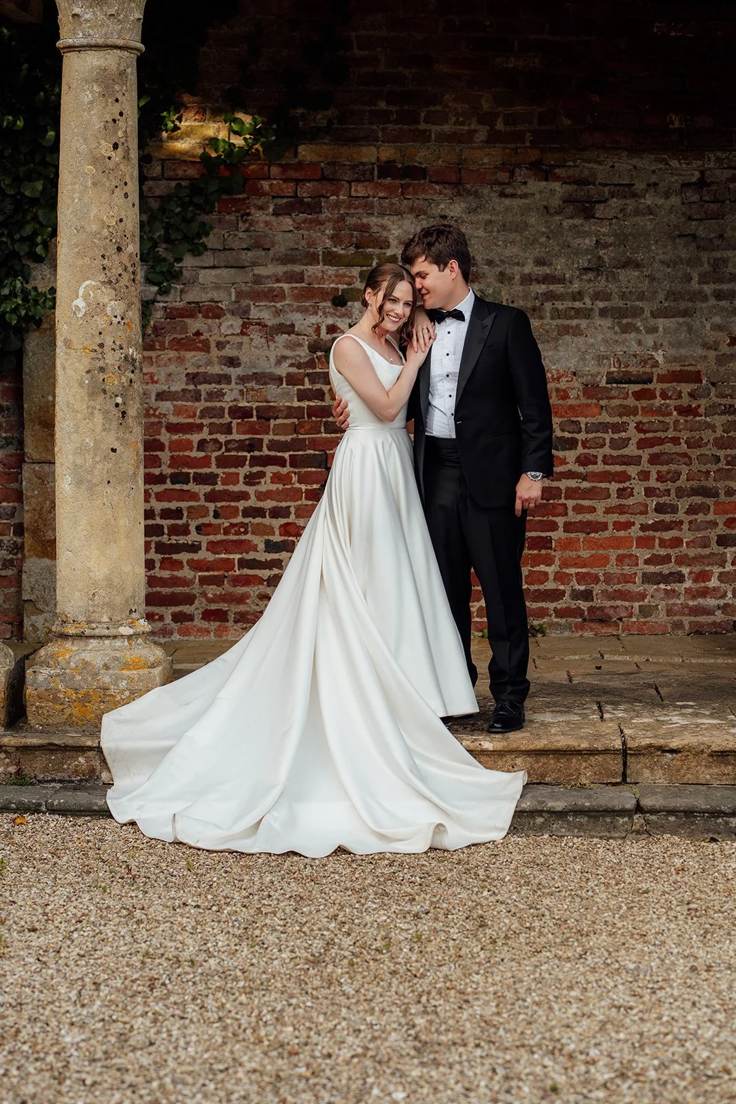 Rachel & Tom stand closely together beside an old brick wall and stone column in the grounds of Brympton House in Somerset, photographed by Just James Weddings.