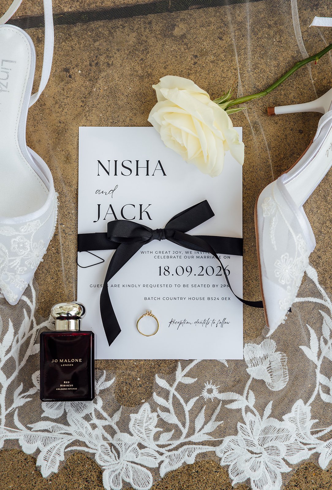 Nisha and Jack’s wedding details at Batch Country House in Somerset, UK, including their invitation, white heels, perfume, engagement ring and a cream rose arranged over lace fabric before the ceremony, all photographed by Just James Weddings.