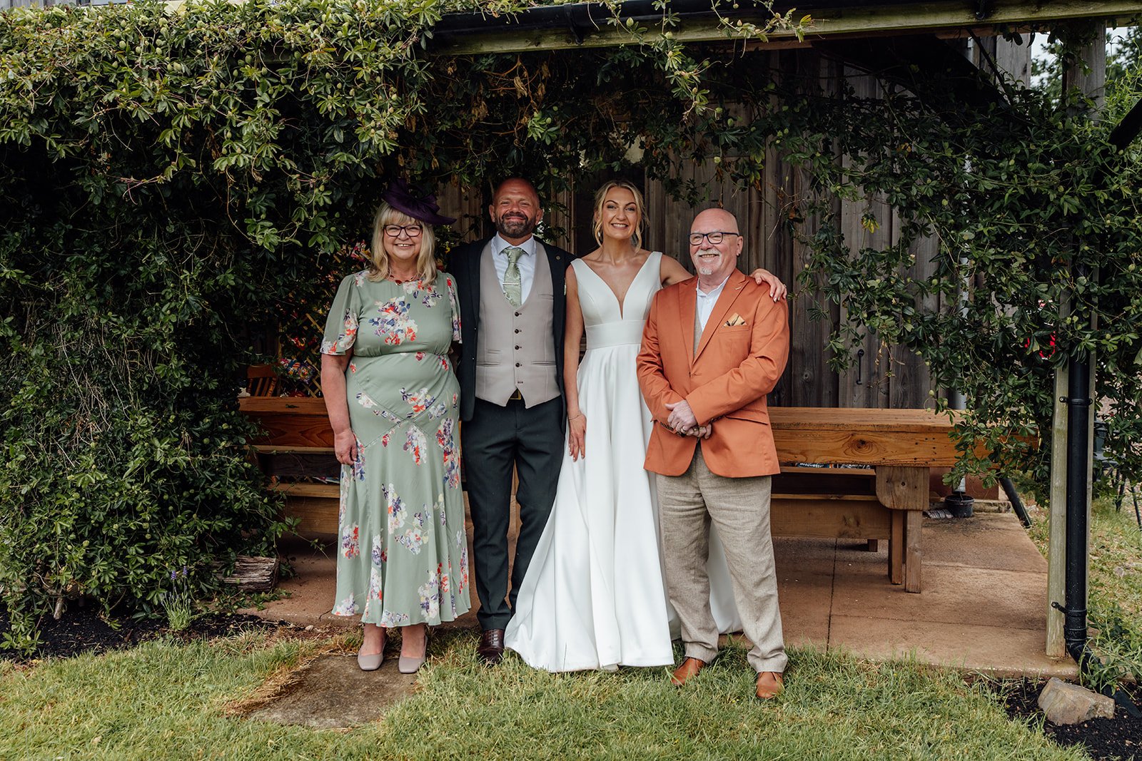 Family portrait of Hannah & Ben at Grays Barn in Devon, UK, standing outdoors with parents beneath leafy greenery and rustic barn wood after the ceremony, captured naturally with warm, relaxed smiles and countryside charm by Just James Weddings.