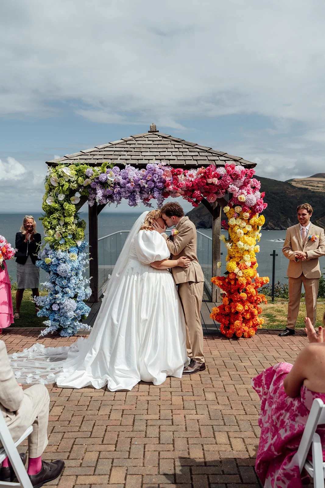 Holly & Alex share their first kiss beneath a colourful floral arch during their outdoor ceremony overlooking the coast at Sandy Cove Hotel in North Devon, photographed by Just James Weddings.
