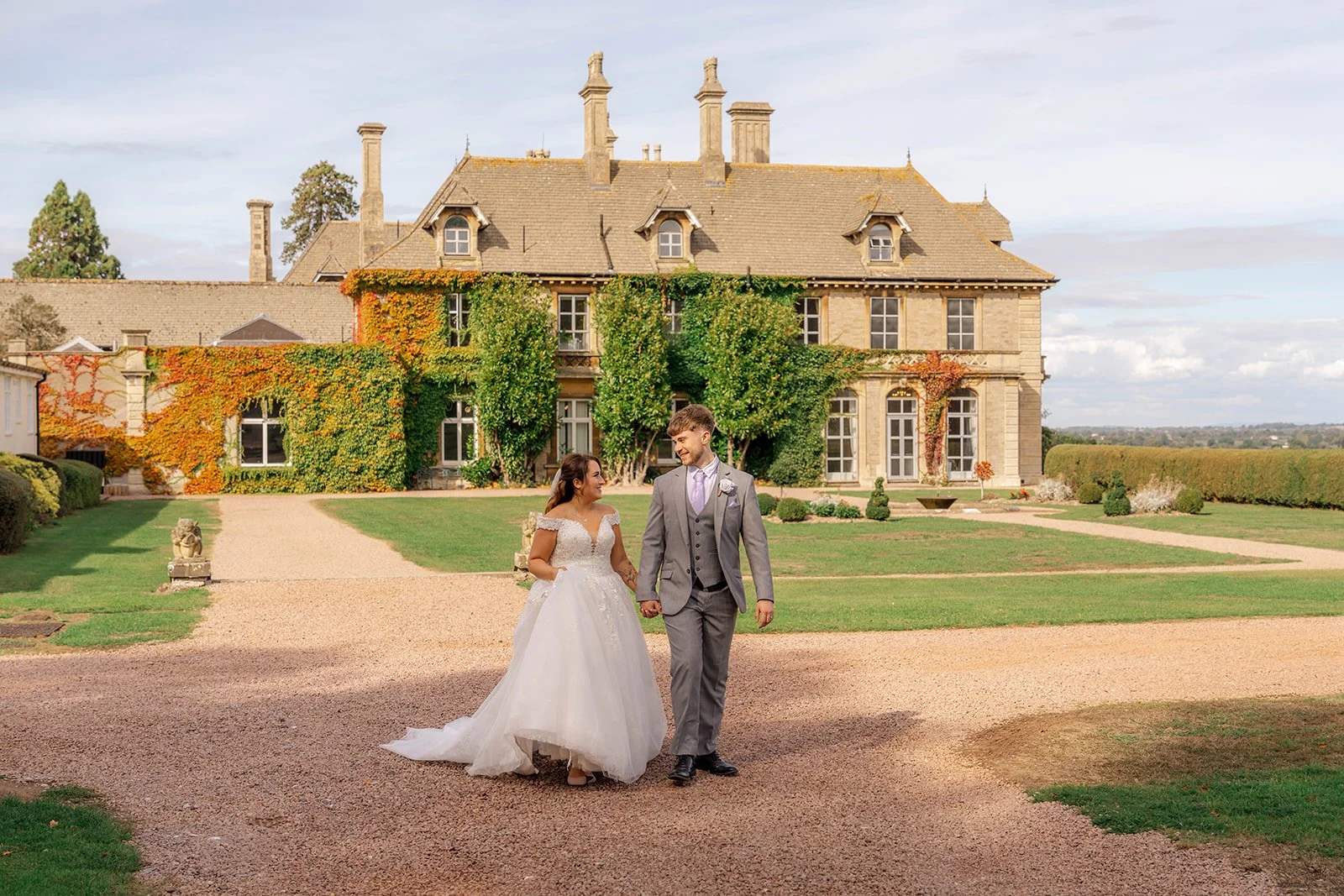 Rhiannon & Anthony walking together outside Eastwood Park in Gloucestershire, with the ivy-covered manor behind them, photographed by Just James Weddings for venue portraits.