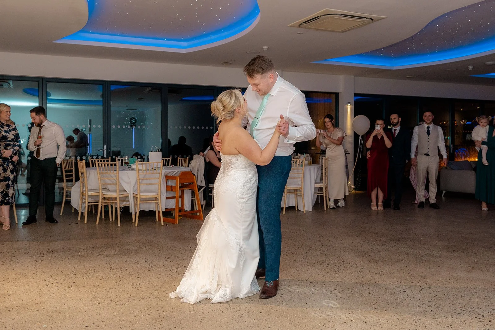 Daisy & Andrew sharing their first dance at Sandy Cove Hotel in North Devon, with guests watching, gold chairs and blue ceiling lights behind them, photographed by Just James Weddings for relaxed coastal wedding reception photography moments.