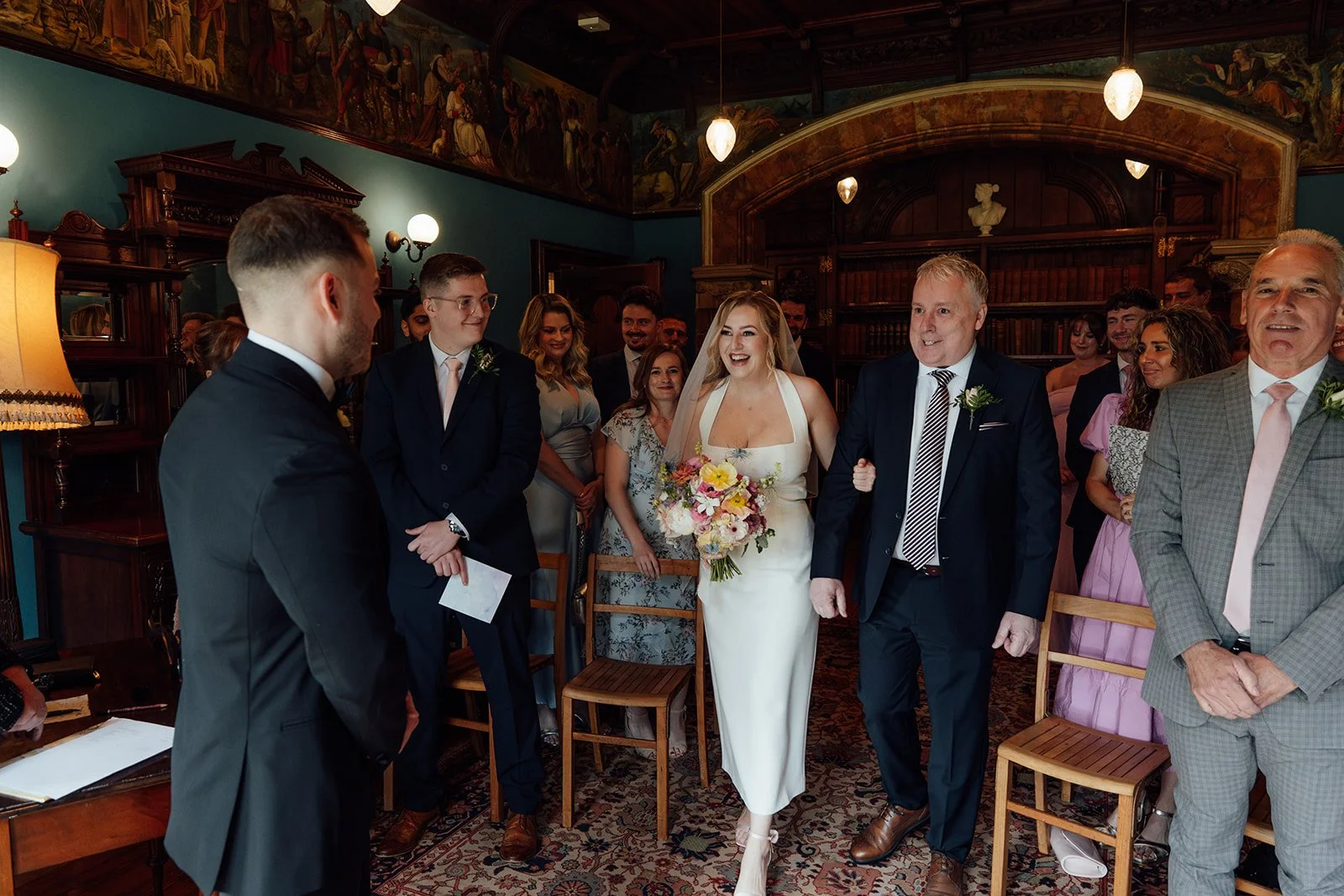 Bryony walking down the aisle with her dad during Bryony & Max’s wedding ceremony at Insole Court in Cardiff, surrounded by smiling guests, rich blue walls, warm lighting, patterned carpet and the venue’s historic interiors on their special day.