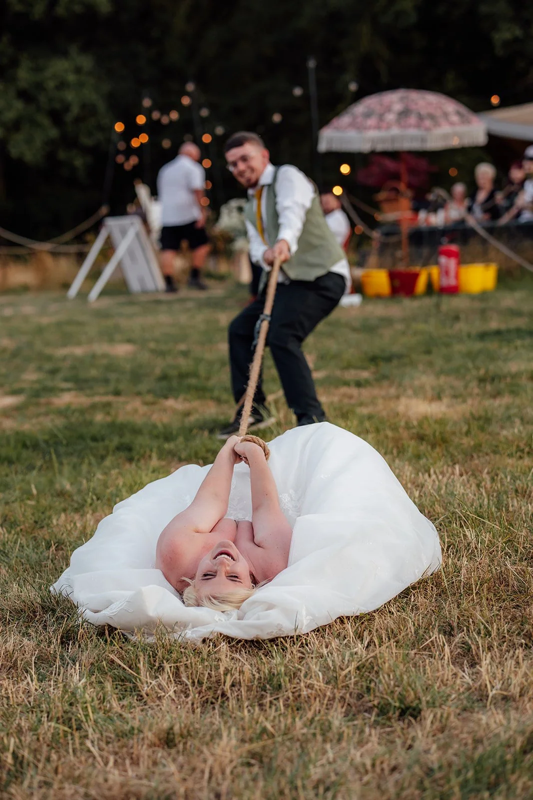 Lucy laughing on the grass while Matthew pulls the rope during a playful wedding tug of war at Curated Canvas at Noah’s Pond in Somerset, photographed by Just James Weddings.