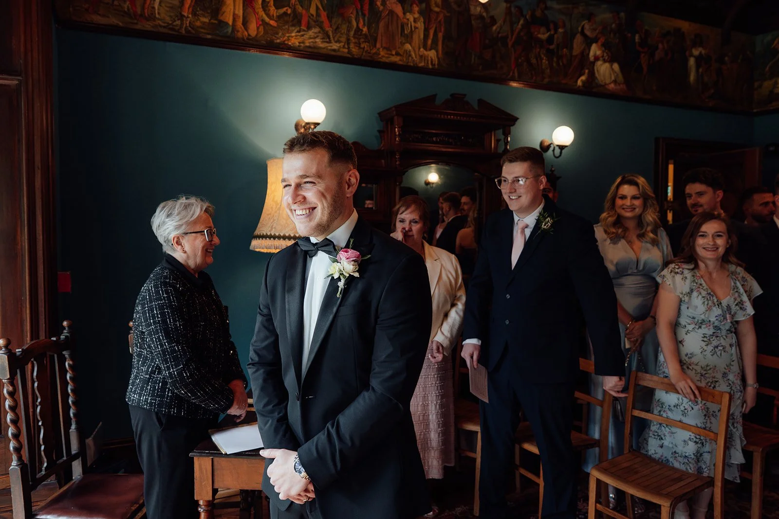 Max smiling at the front of the ceremony room at Insole Court in Cardiff as guests gather behind him, capturing a joyful, candid moment before Bryony walks down the aisle during Bryony & Max’s intimate wedding day in the historic room space.