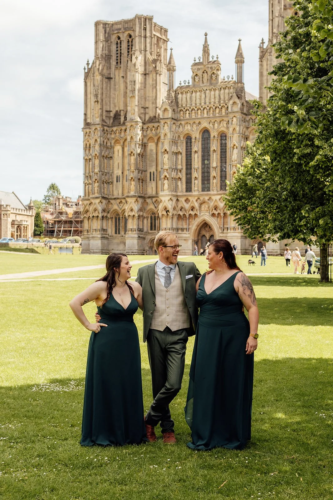 Charlotte & Jack wedding party portrait at Bishop’s Palace in Somerset, UK, with the groom laughing between two bridesmaids in dark green dresses on the lawn, with Wells Cathedral rising behind them on a sunny wedding day full of relaxed energy.