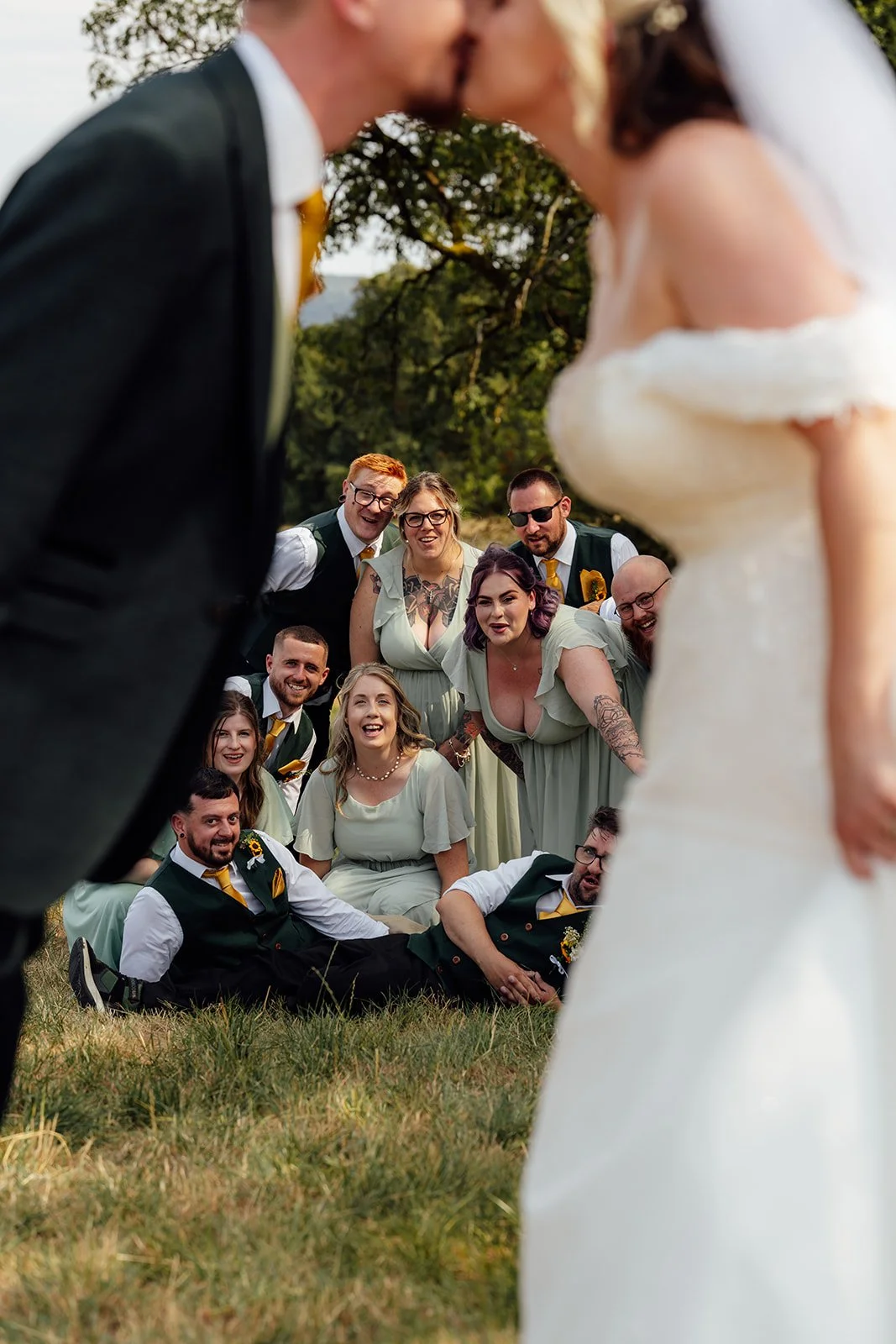 Lucy and Matthew kissing in the foreground while their wedding party reacts playfully behind them at Curated Canvas at Noah’s Pond in Somerset, photographed by Just James Weddings.