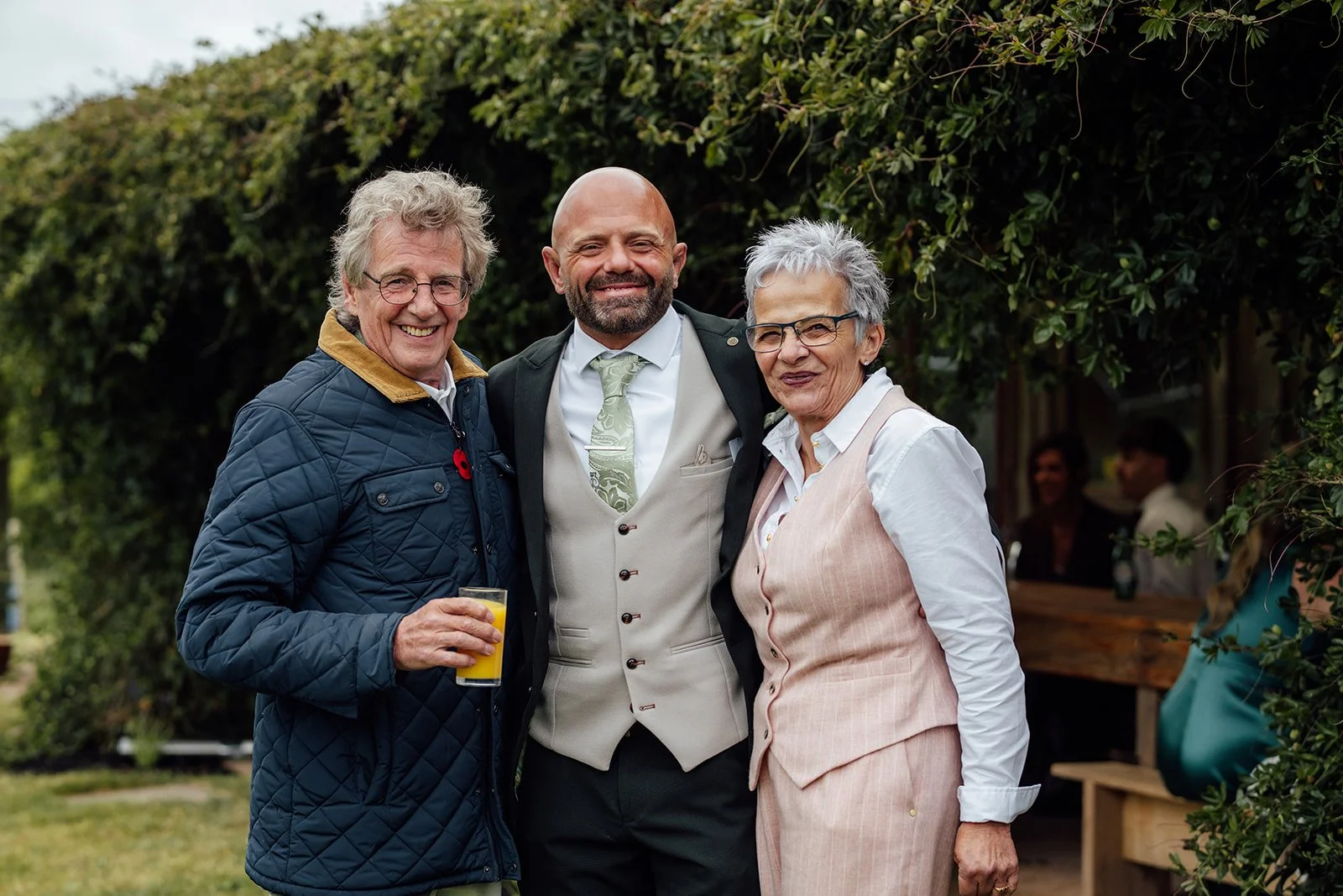 Relaxed family portrait of Ben with two guests at Grays Barn in Devon, UK, smiling together beneath leafy greenery after the ceremony, with countryside charm, drinks and warm candid energy captured naturally by Just James Weddings outdoors.