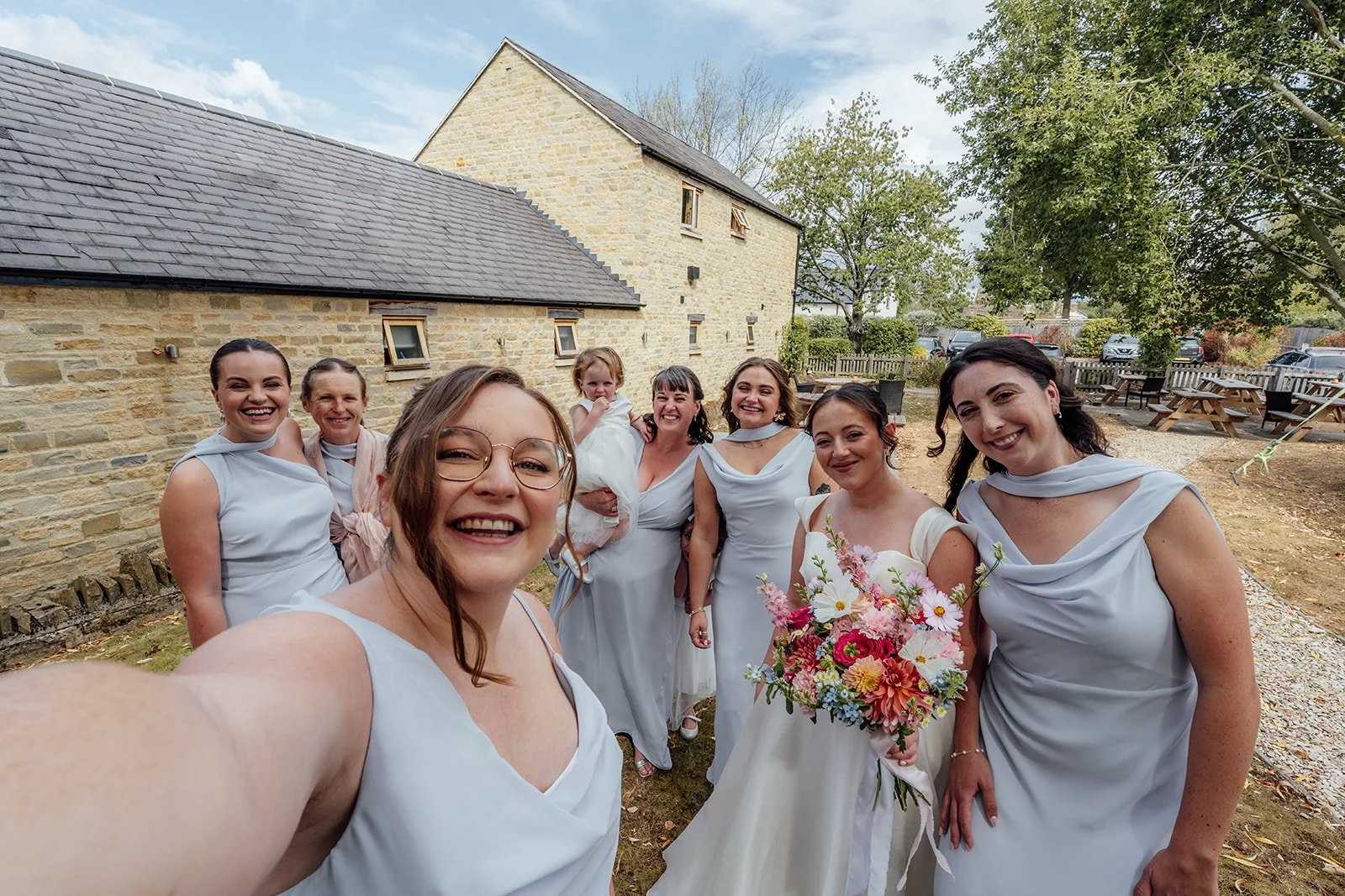 Harriet poses with her bridesmaids and flower girl for a cheerful outdoor selfie beside the stone buildings at Dovecote Events in Oxfordshire, photographed by Just James Weddings.