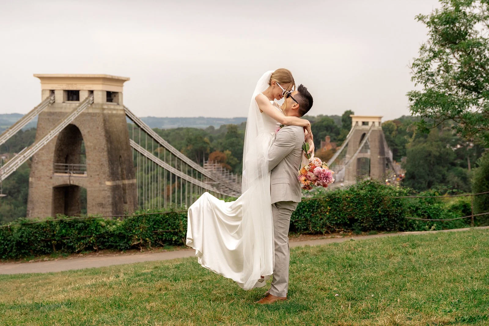 Brian & Josie kissing with the bride lifted in front of Clifton Suspension Bridge at Clifton Observatory in Somerset, photographed by Just James Weddings for fun romantic wedding portraits.