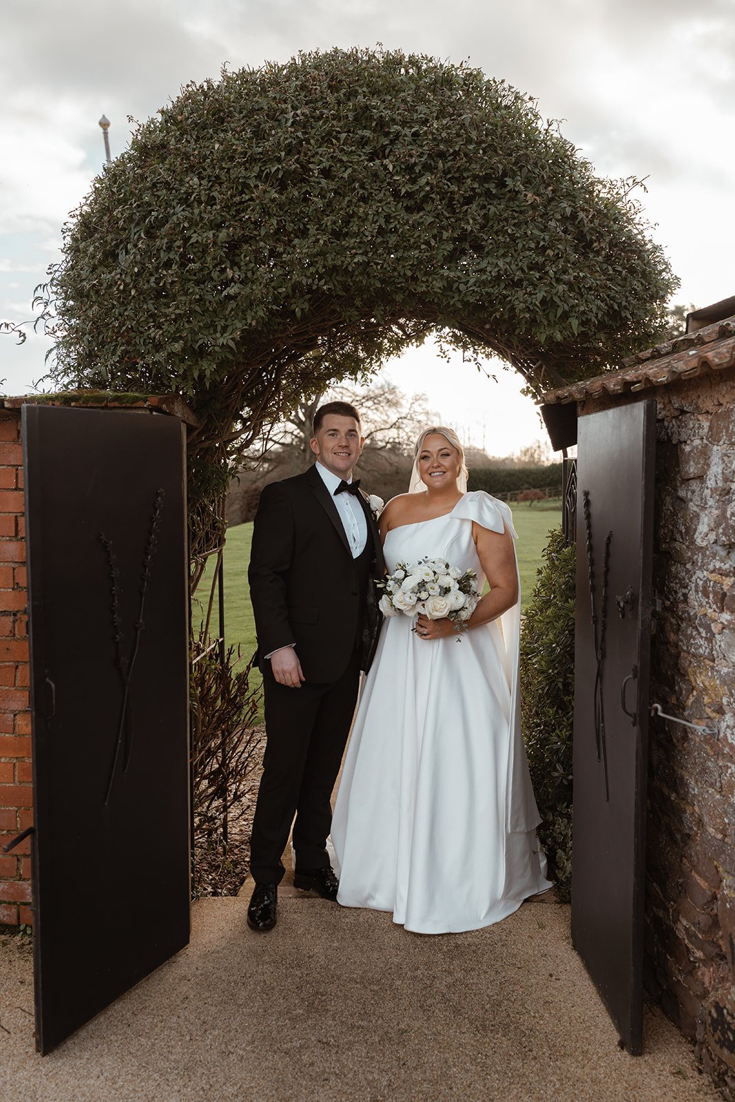 Jessie & Jay stand together beneath a leafy garden arch at Upton Barn & Walled Garden in Devon, UK. The couple smile toward the camera, framed by brick walls and greenery, creating a timeless portrait moment during their wedding day.