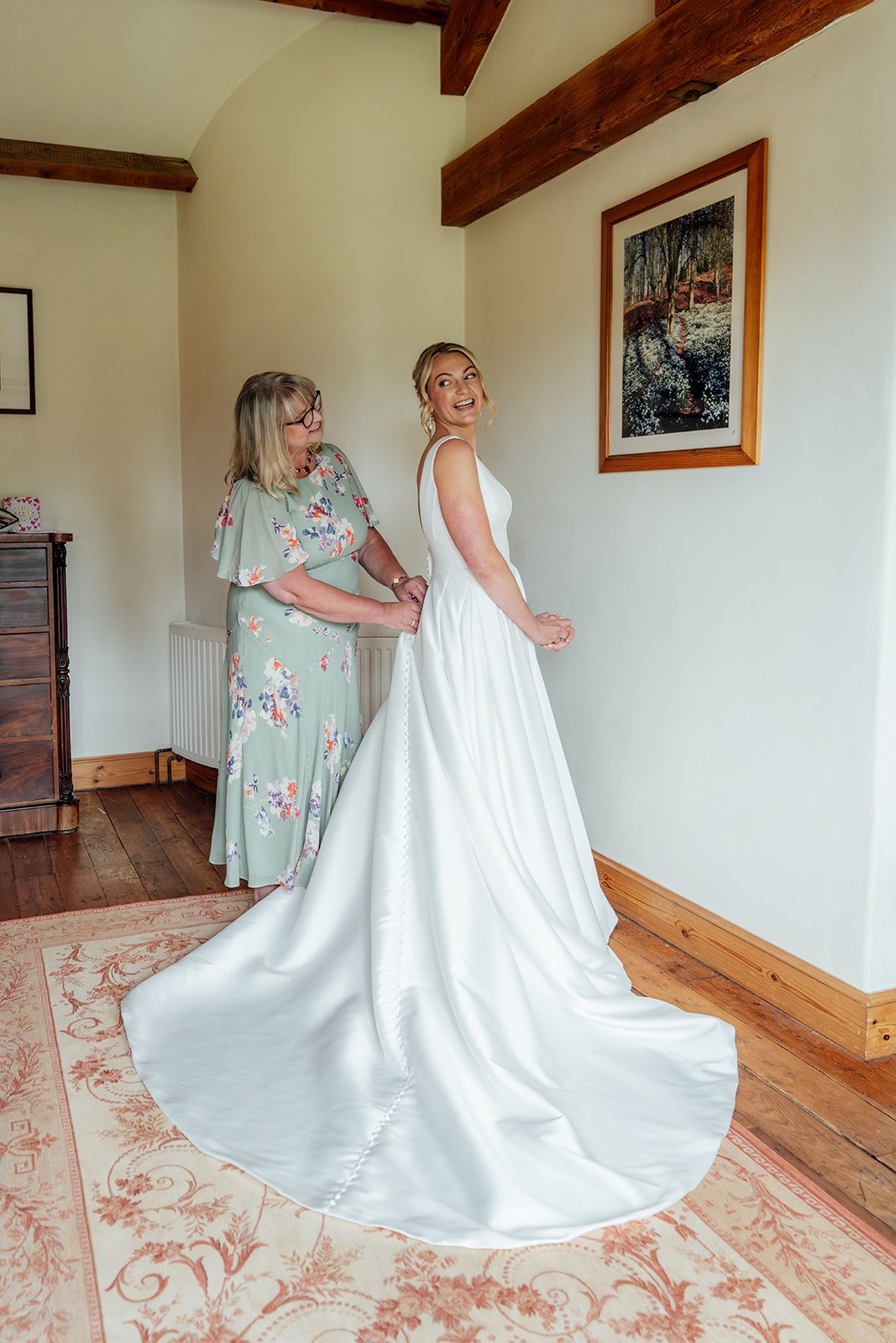 Joyful bridal prep photo of Hannah having her wedding dress buttoned at Grays Barn in Devon, UK, with her mum smiling beside her as the gown’s long train spreads across the wooden floor, warmly photographed beautifully by Just James Weddings.