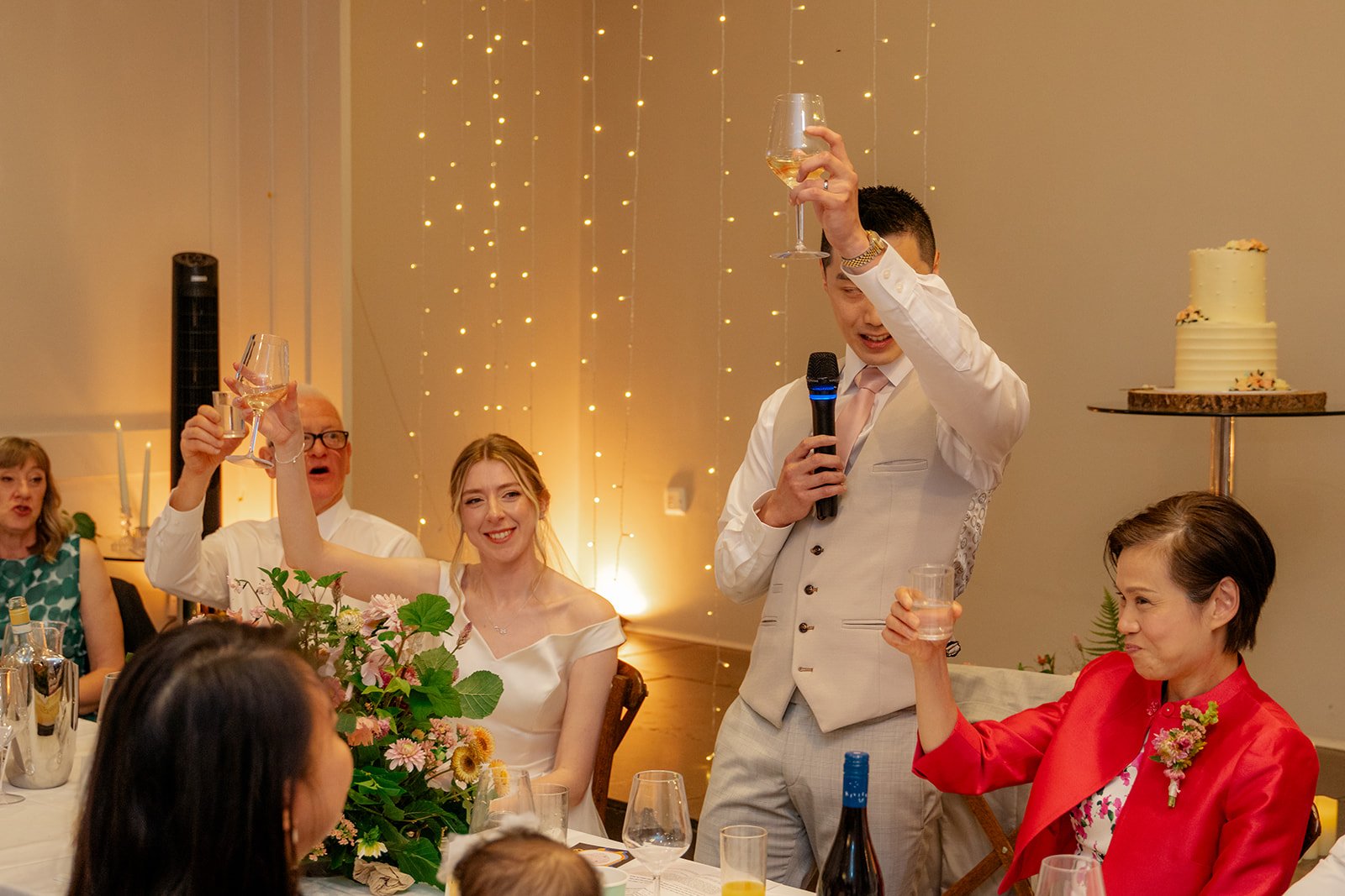 Brian & Josie laughing during wedding speeches at Clifton Observatory in Somerset, with guests raising glasses and fairy lights behind them, photographed by Just James Weddings