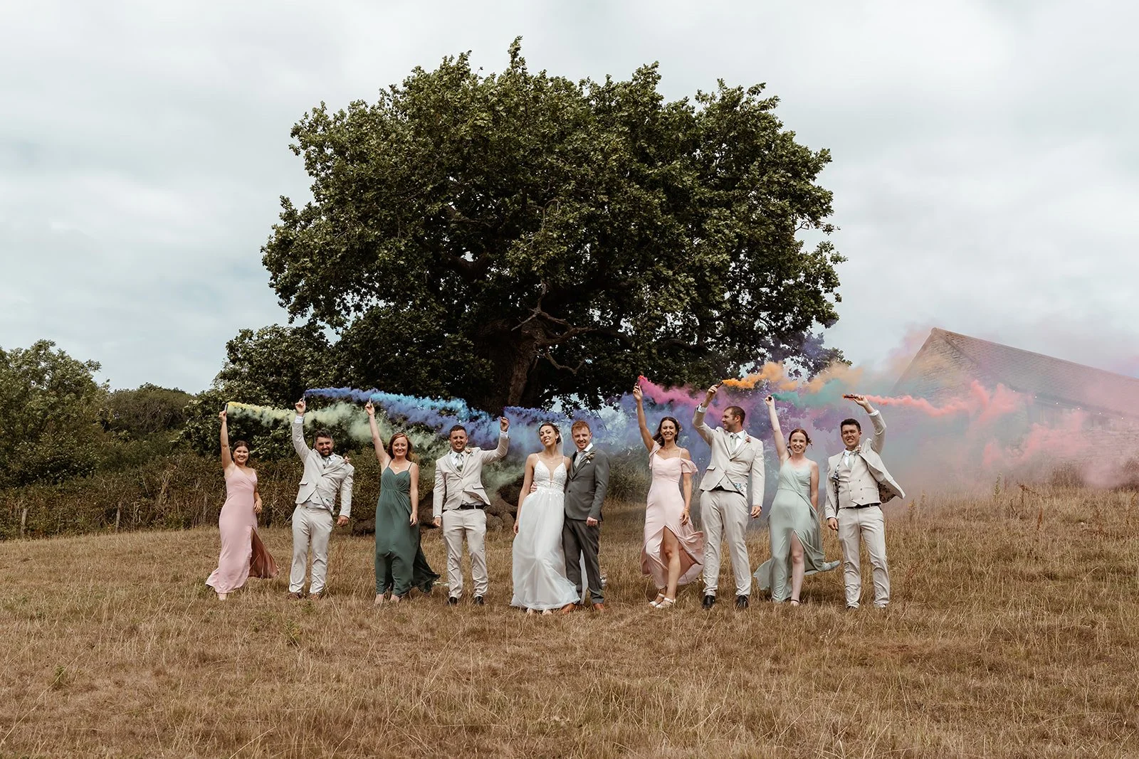 Madeleine &amp; Izaac at Folly Farm Centre celebrating with their wedding party in a wide outdoor field, everyone holding colourful smoke bombs that create a vibrant rainbow trail behind them as they stand together in front of a large tree.