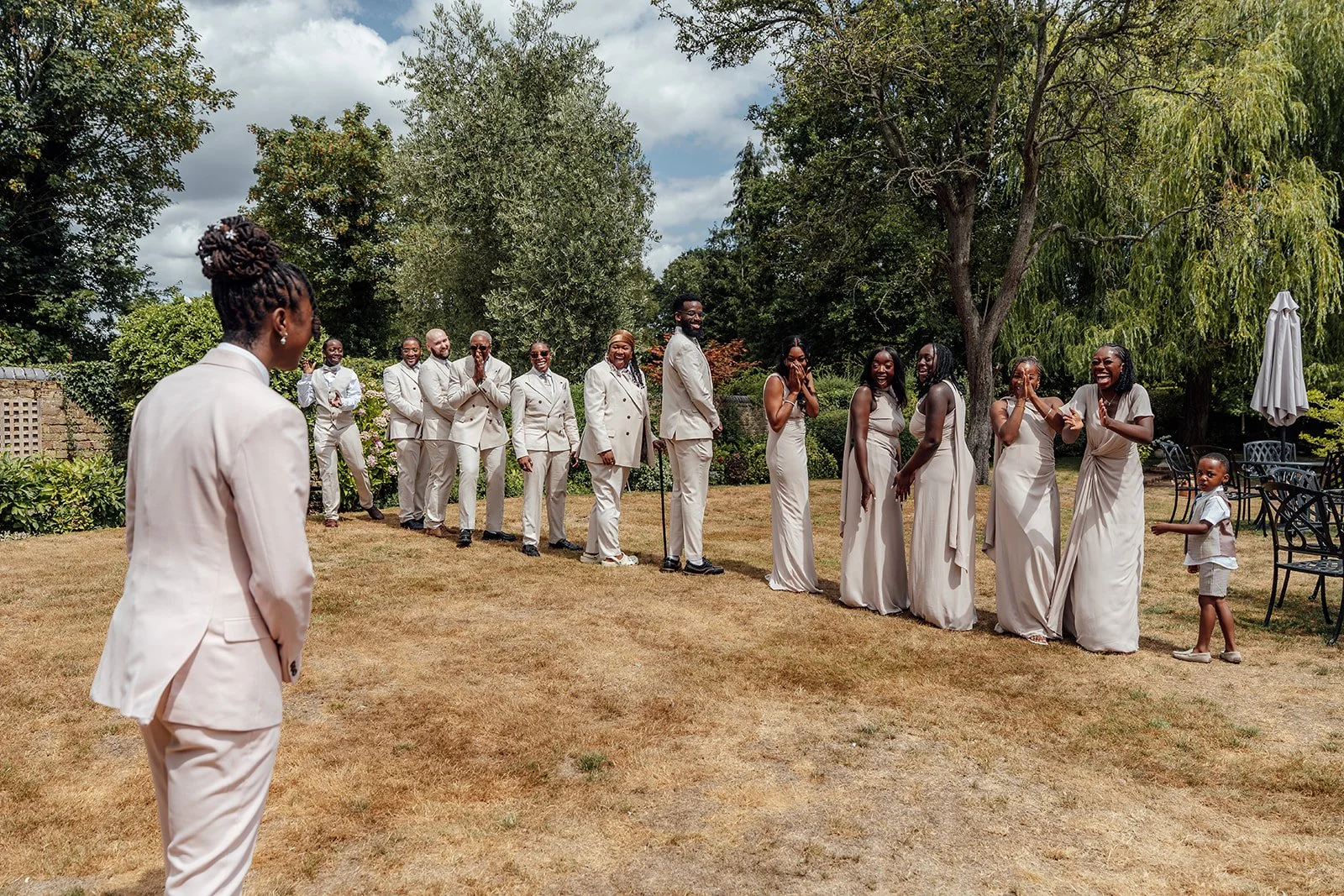Kasey approaching the wedding party as friends react with excitement during a playful group photo in the gardens at Oaks Farm Weddings in Surrey.