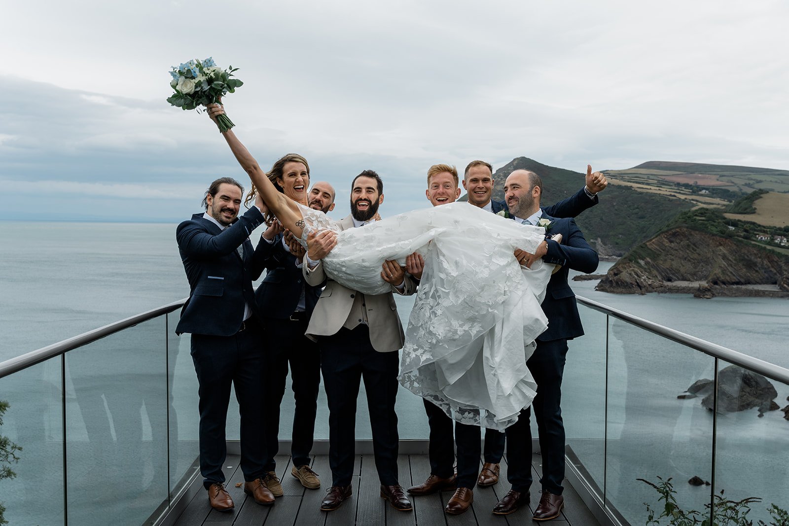 Jazz & Peter’s groomsmen lifting the bride on the balcony at Sandy Cove Hotel in North Devon, with sea and cliff views behind them, captured by Just James Weddings in a fun documentary wedding photography style after their coastal ceremony.