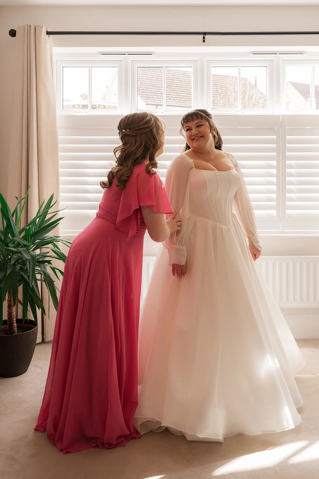 Emily & Josh wedding morning portrait at Coombe Lodge Blagdon, Somerset, UK, showing the bride in her gown smiling with a woman in a bright pink dress by the window. Soft natural light and a calm getting-ready moment capture the joy of the day.