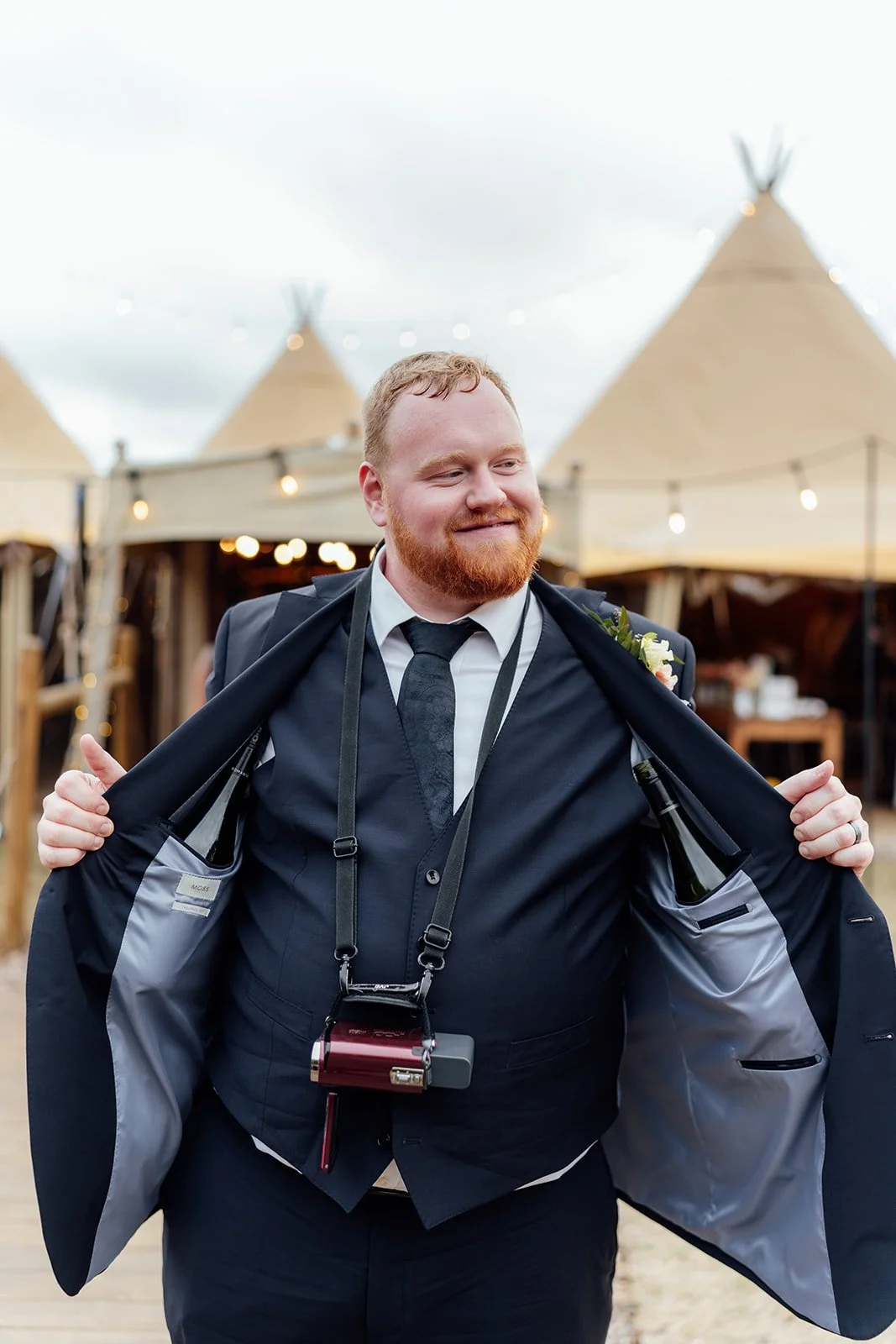 Wedding guest in a navy suit opens his jacket to reveal hidden bottles and a camera during the reception at Dovecote Events in Oxfordshire, photographed by Just James Weddings.