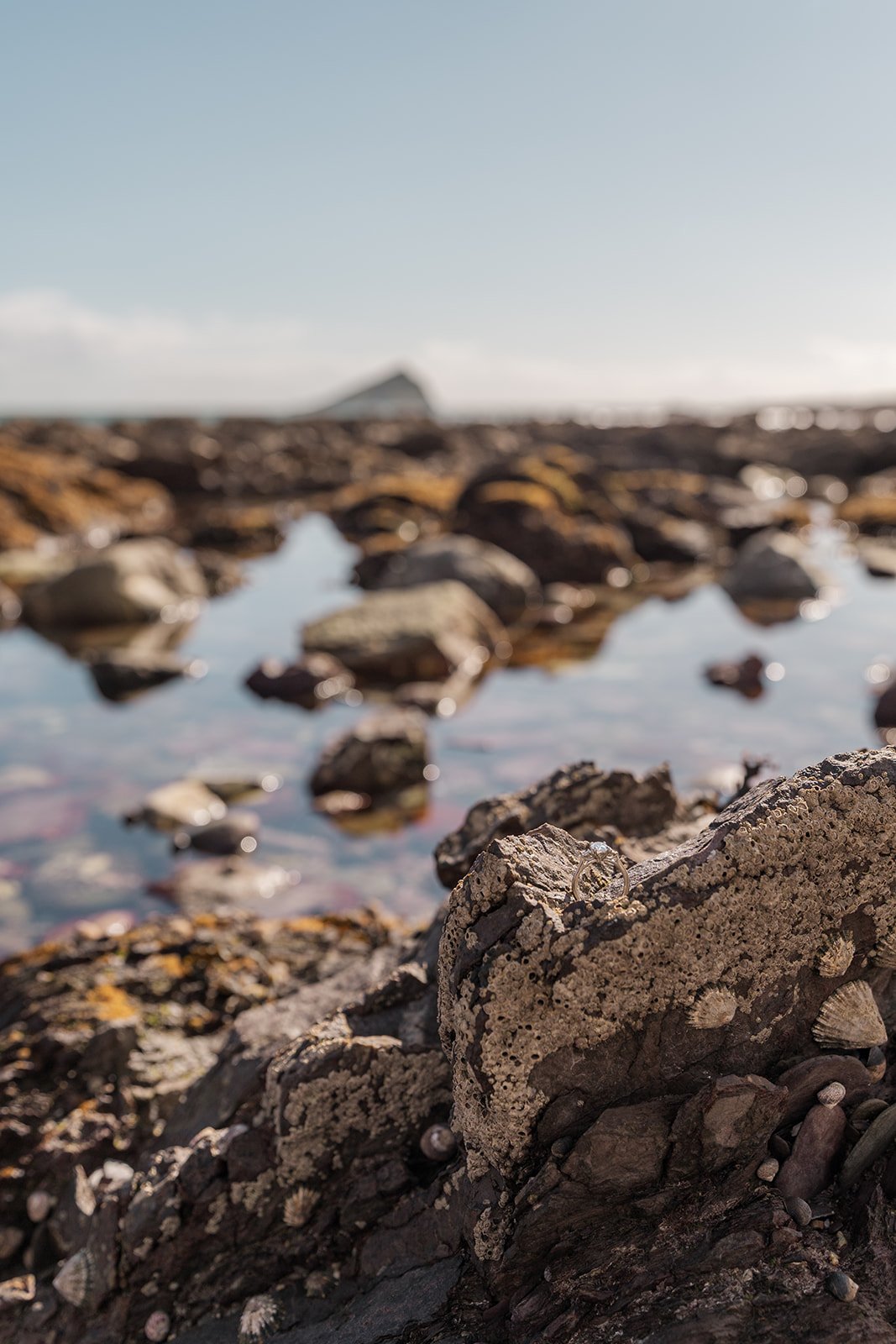 Katie and Ben’s engagement ring resting on a textured seaside rock beside clear tide pools and coastal stones, with the Somerset shoreline softly blurred behind during a relaxed beach engagement photo session captured by Just James Weddings.