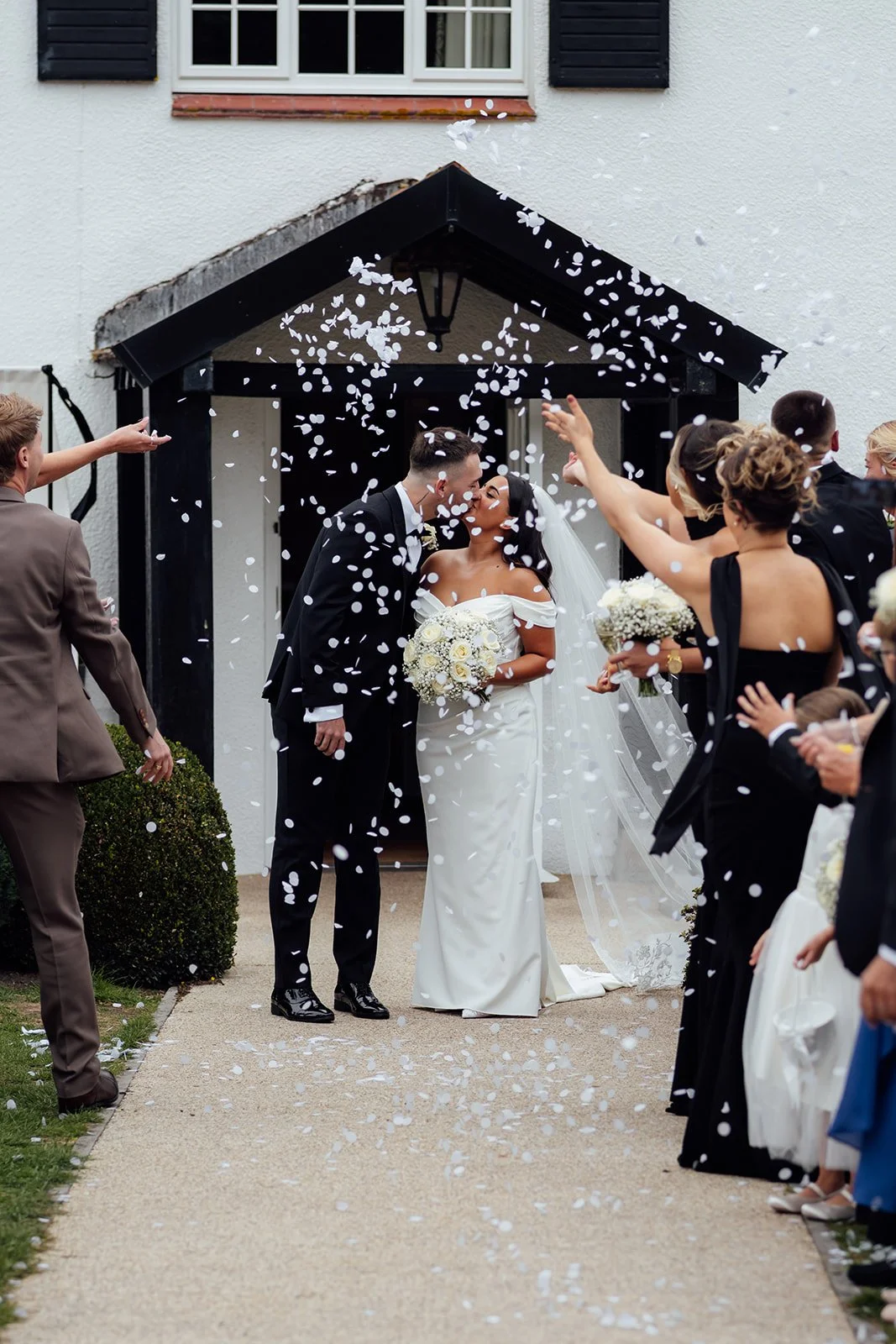 Nisha and Jack share a kiss beneath falling confetti outside Batch Country House in Somerset, UK, surrounded by cheering wedding guests as Nisha holds her white floral bouquet and her veil falls behind her, photographed by Just James Weddings.