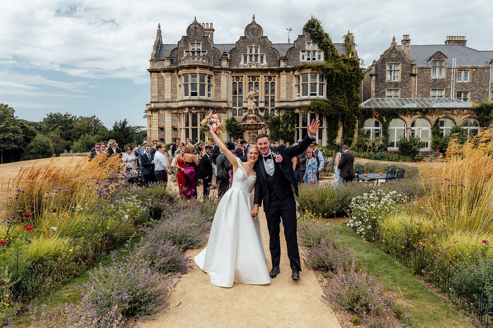 Caitlin and Kevin celebrating together in the gardens at Clevedon Hall in Somerset, raising their arms with the historic wedding venue and guests behind them.