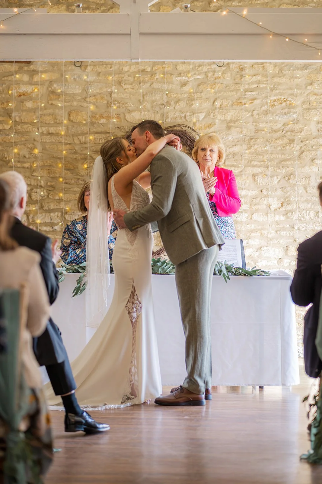Anna and Tom share their first kiss during their wedding ceremony at Winkworth Farm in the Cotswolds, UK. The indoor barn setting, stone wall backdrop and warm fairy lights frame a joyful just-married moment in this romantic wedding photograph.
