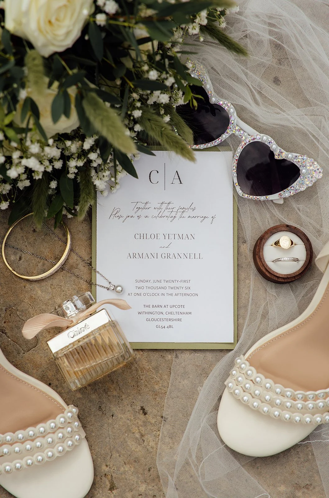 Wedding detail flat lay from Chloe & Armani’s day at The Barn at Upcote in the Cotswolds, featuring their invitation, heart-shaped sunglasses, perfume, rings and pearl wedding shoes.