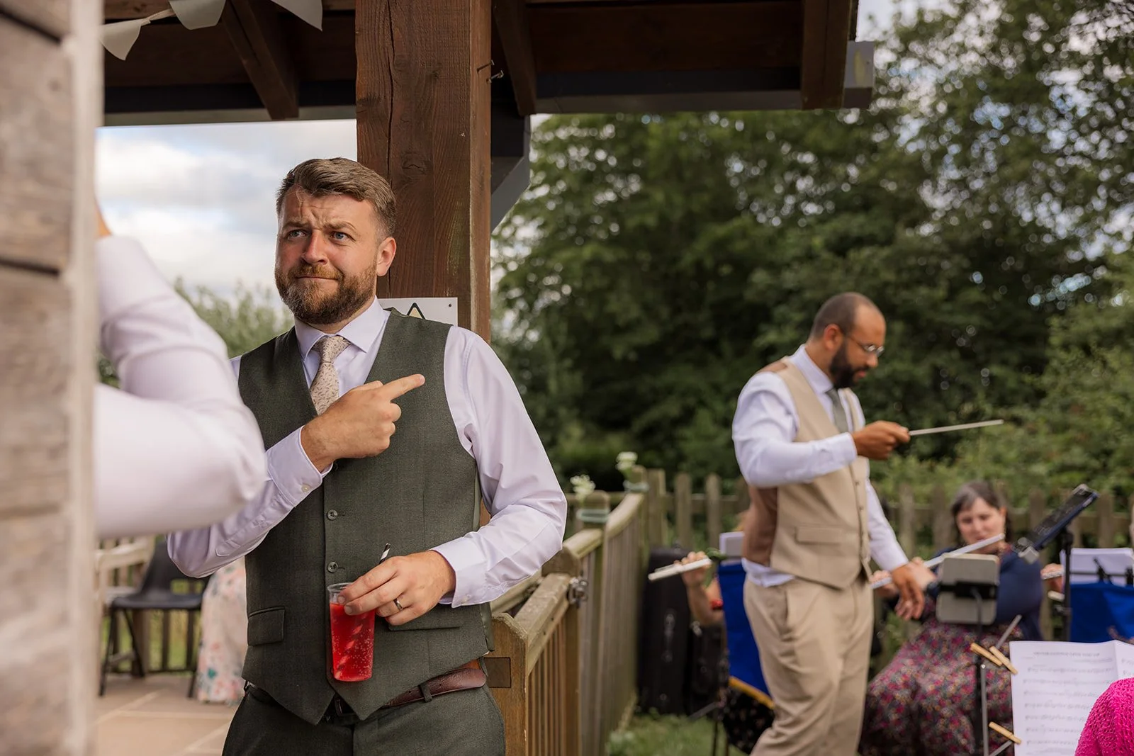 Jack holds a drink and gestures playfully while Aaron stands beside the live band during their reception at Rock Village Hall in Worcestershire, photographed by Just James Weddings.