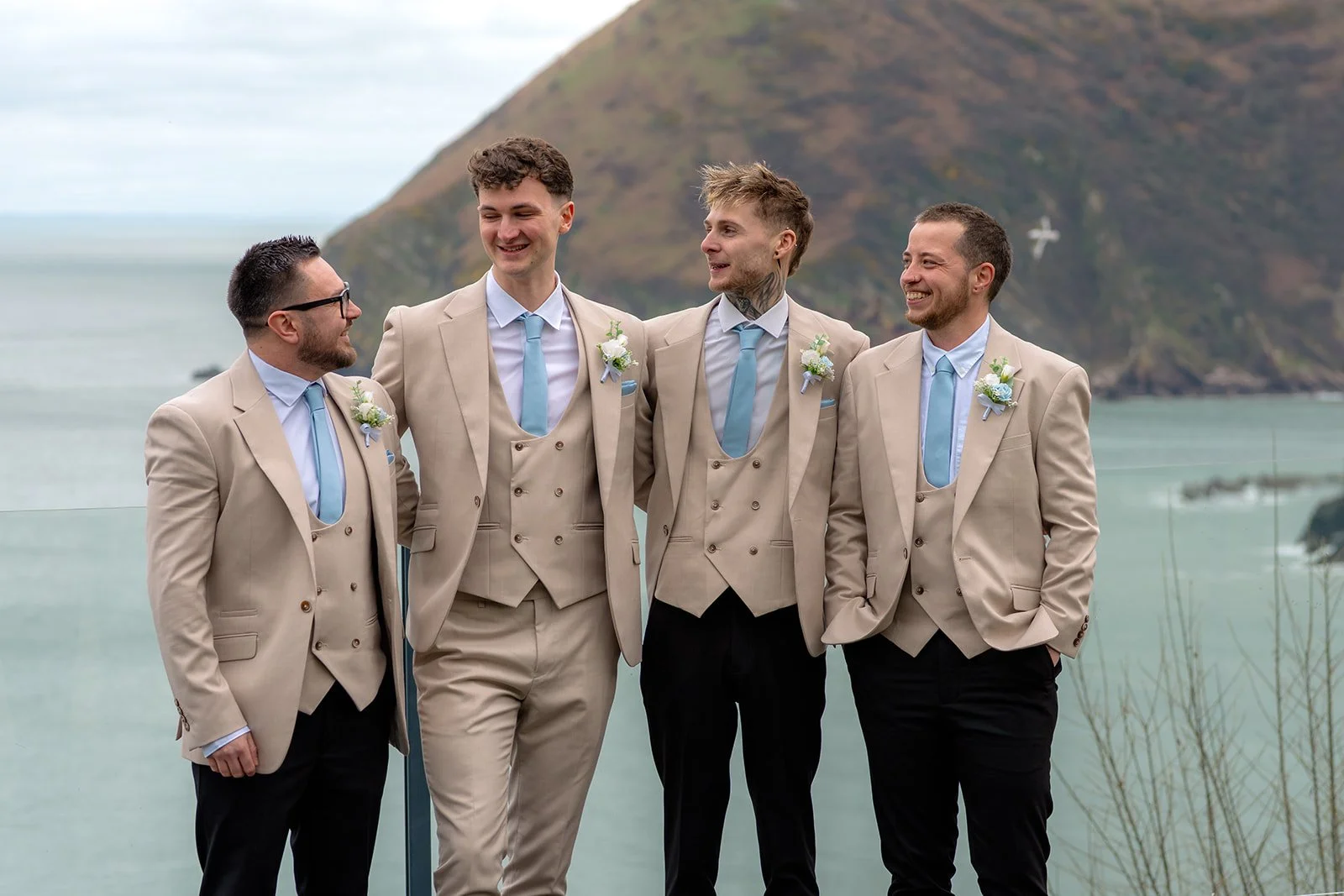 Groom Cameron with groomsmen in beige suits and blue ties overlooking the North Devon coastline during Hannah & Cameron’s wedding at Sandy Cove Hotel. Relaxed groom portraits with sea views and cliffs on their coastal wedding day celebration.