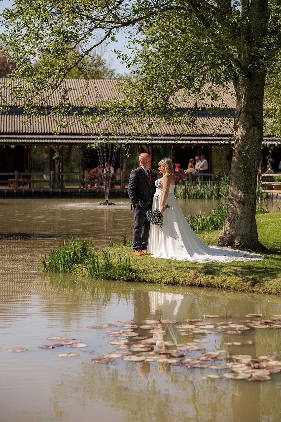 Debbie and Nigel stand beside the lake at Yarlington Barn in Somerset for a quiet couple portrait, with the barn reflected in the water, trees overhead, and sunny countryside gardens.