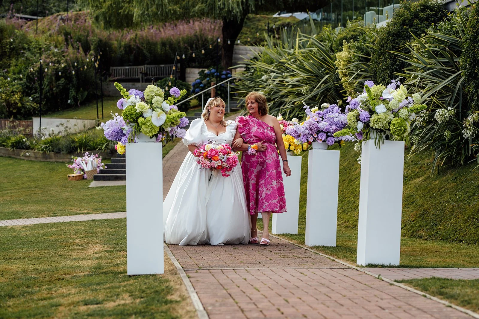 Holly walks towards the outdoor ceremony with her mum beside colourful floral displays at Sandy Cove Hotel in North Devon, photographed by Just James Weddings.