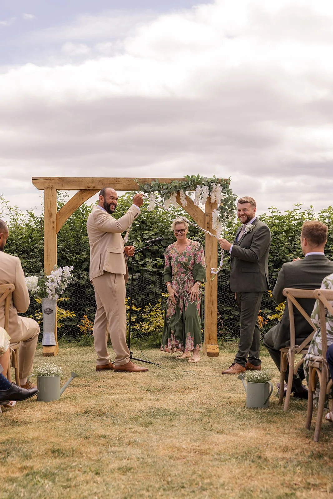 Aaron & Jack smile while holding a colourful ribbon together during their outdoor wedding ceremony beneath a floral wooden arch at Rock Village Hall in Worcestershire, photographed by Just James Weddings.