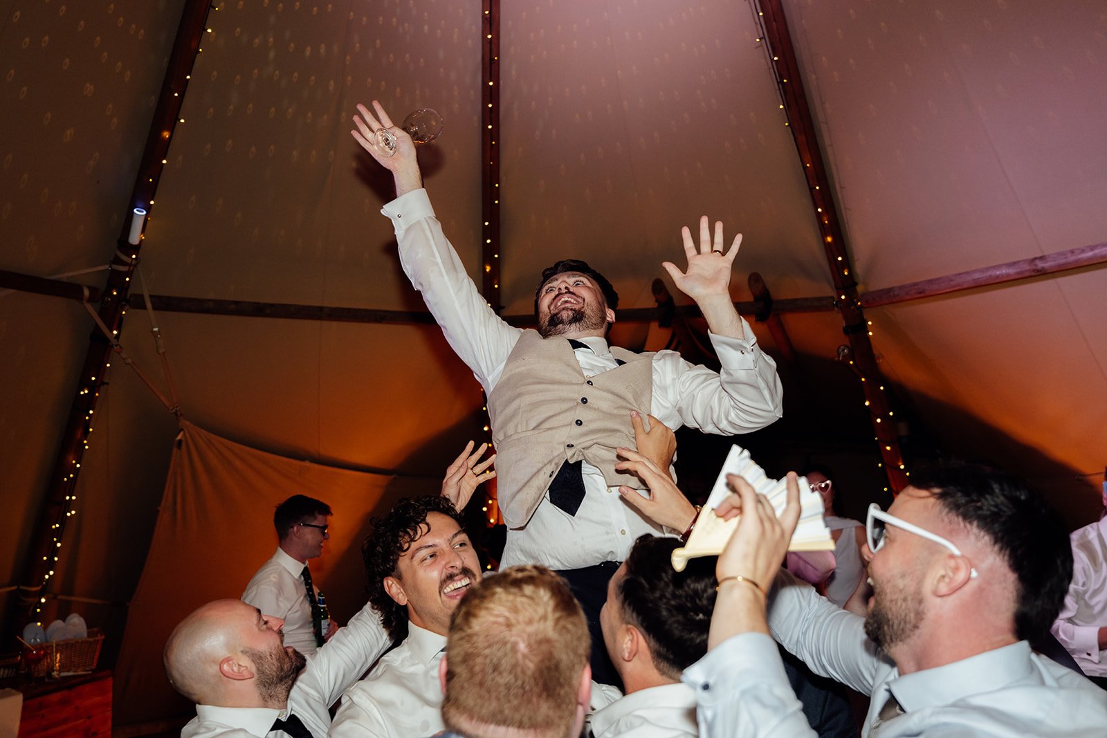 Simon is lifted into the air by friends while holding a drink during the lively evening reception at Dovecote Events in Oxfordshire, photographed by Just James Weddings.