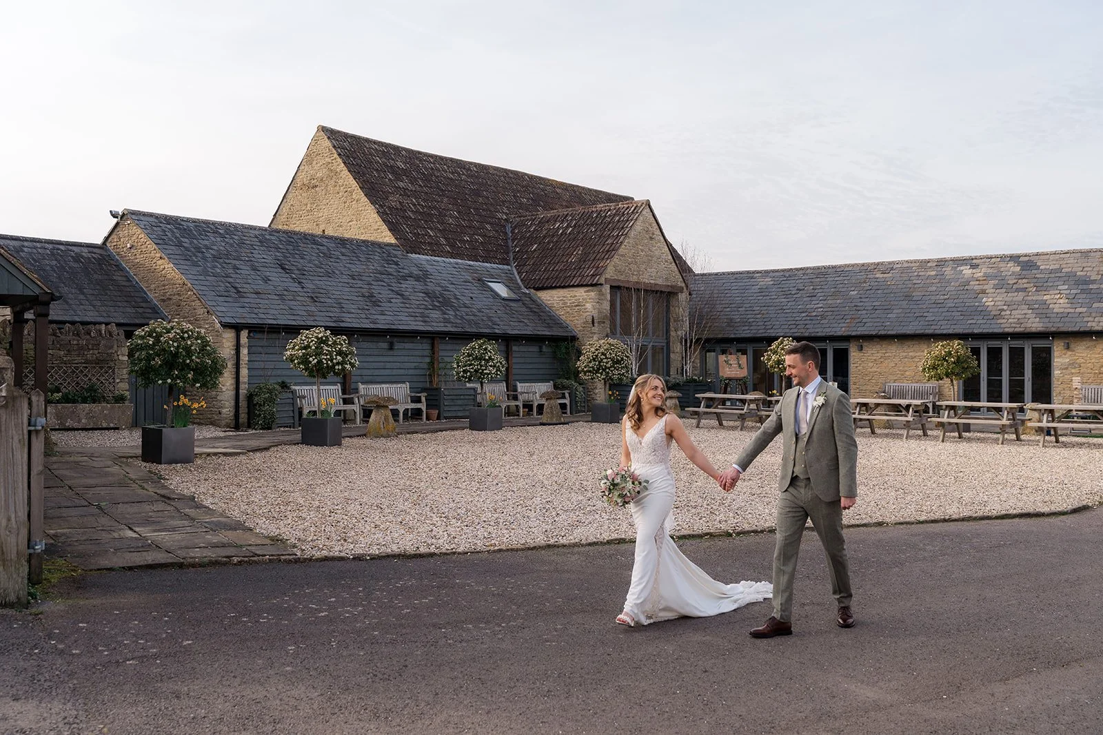 Anna and Tom walk hand in hand across the courtyard at Winkworth Farm in the Cotswolds, UK, during their wedding portraits. This wide outdoor image captures the stone barn venue, relaxed newlywed energy and natural Cotswolds wedding photography.